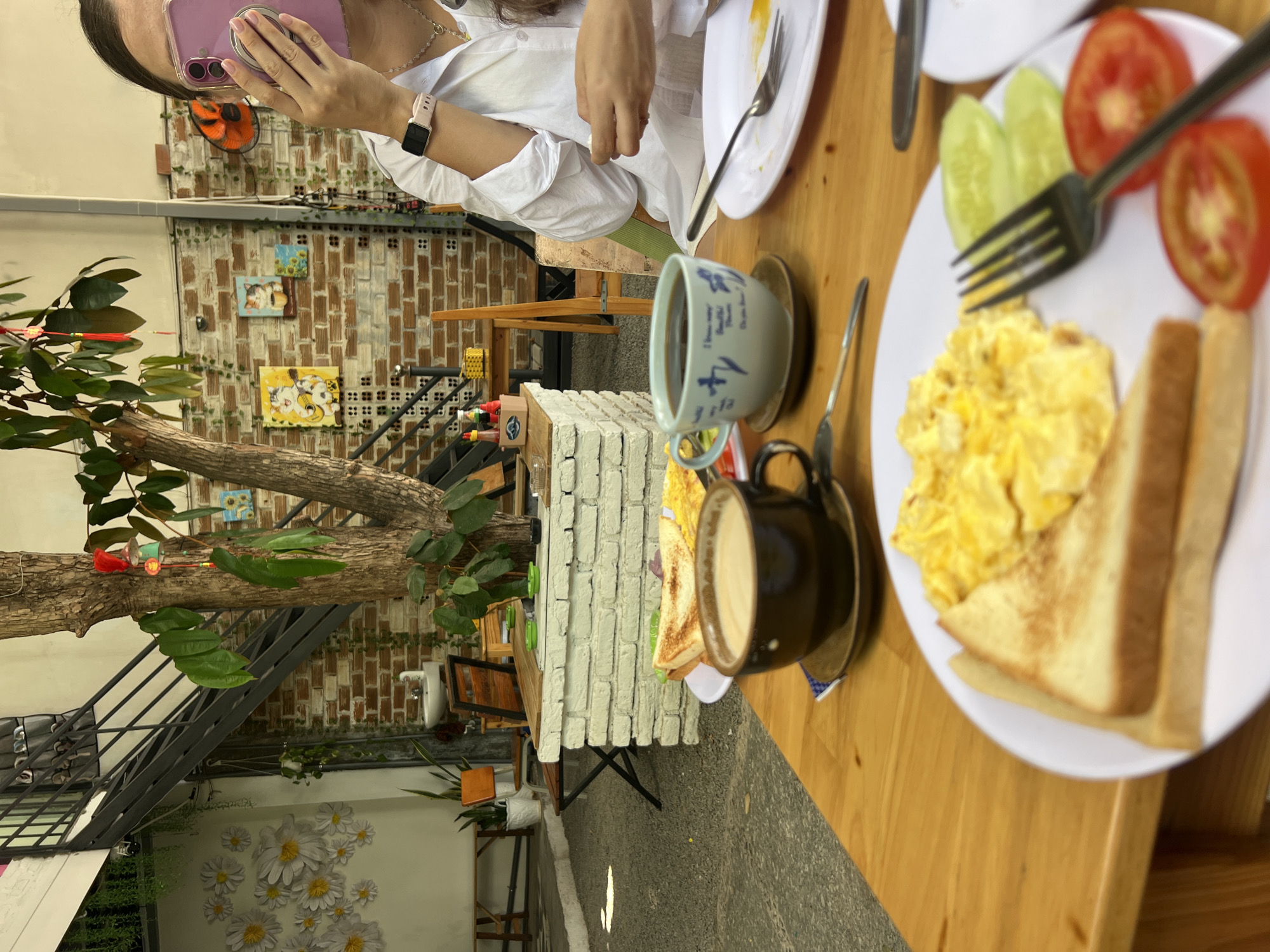 A person with a phone and pink case is wearing a white long-sleeved shirt and sitting at a wooden table. On the table are a plate of scrambled eggs with toast, sliced cucumbers, and tomatoes, and a fork is piercing the tomatoes. Also, there is a black cup and a blue and white cup, both containing liquid. Part of another white plate is visible in the top right corner.
The background shows a brick wall with small framed pictures and a staircase leading to an upper level. A tree is growing through an opening in the floor. There are also white flowers and green plants near the stairs. The location is in the city of Bắc Nha Trang, Vietnam.