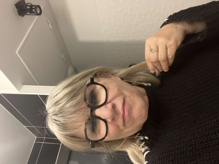 A woman with blond hair and black glasses faces the camera. She is wearing a dark sweater and earrings. Her right hand is raised with a ring on her finger. The background features a white tiled wall with a square pattern, a black tiled wall, a white door, and a black security camera. The image appears to have been rotated counter-clockwise.