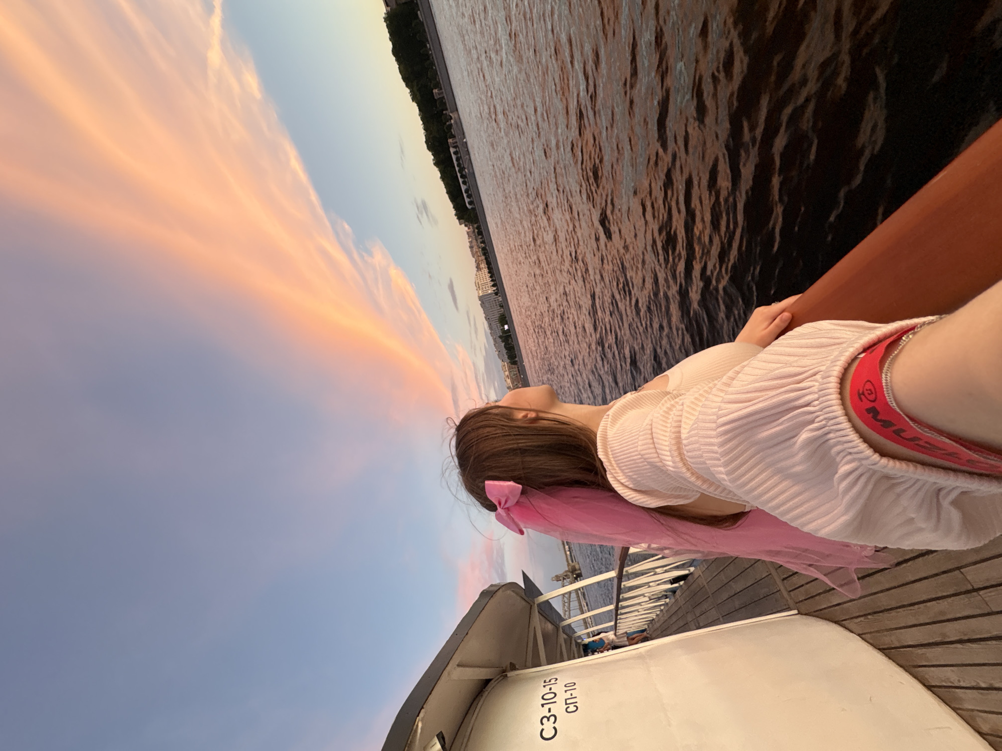 The image shows a young woman on a boat, facing away from the camera and looking at a sunset over a body of water. The sky is filled with streaks of pink and orange clouds. The woman has long brown hair adorned with a pink bow and veil. She is wearing a light-colored, ribbed top. On the side of the boat, there is writing that reads "СЗ-10-15" and "СП-10". The boat is docked on a body of water, likely a river or canal, with buildings and trees visible on the opposite bank. The scene appears to be set in Saint Petersburg, Russia, given the Cyrillic writing and the architectural style of the buildings.