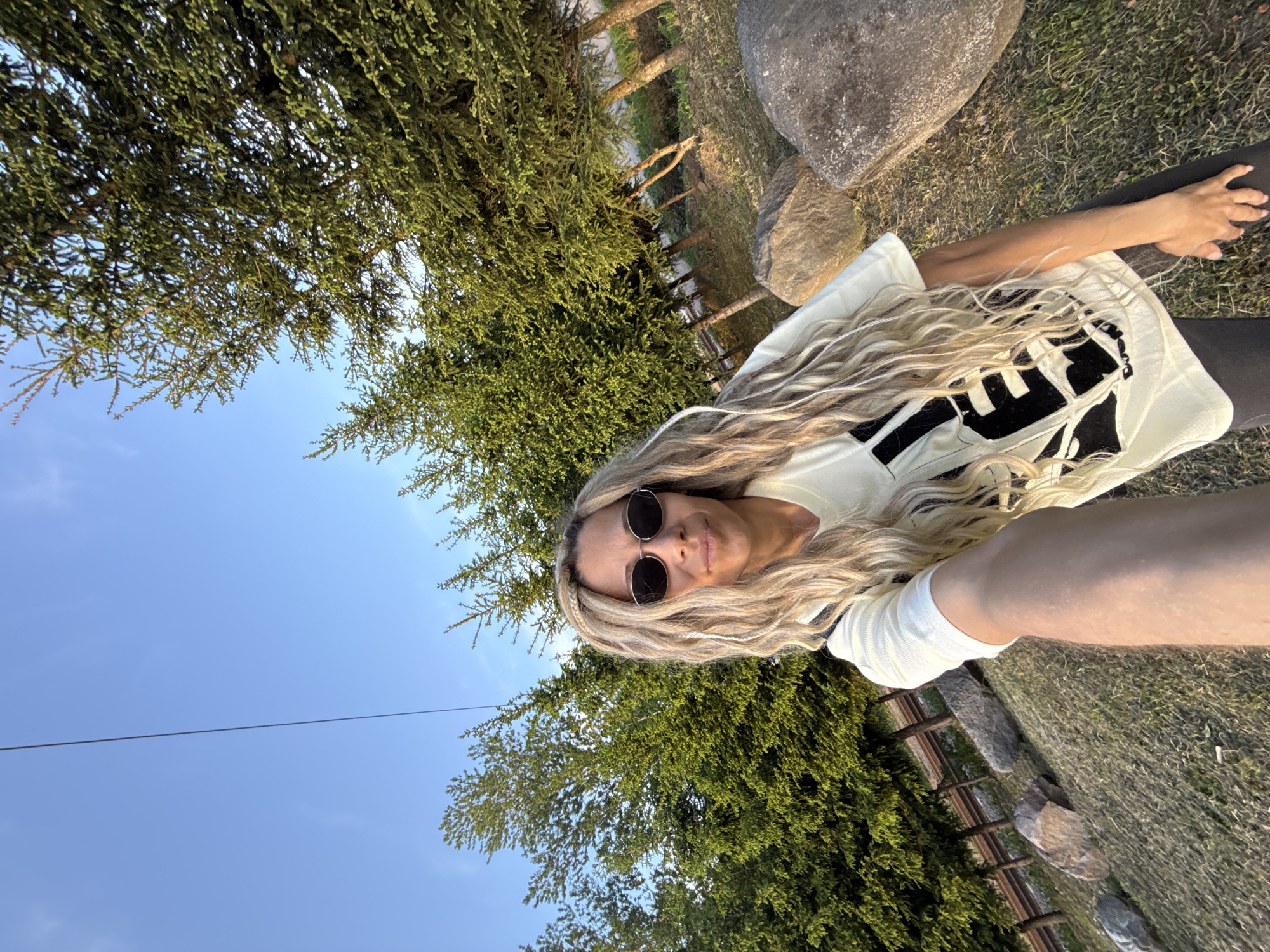 A selfie shot from a low angle shows a blonde-haired woman wearing sunglasses, a light-colored t-shirt with black graphics, and dark leggings. She is positioned on grass, next to a large rock and a wooden fence. Green trees are visible in the background, along with a patch of blue sky. The location is Rusanovo, Russia.