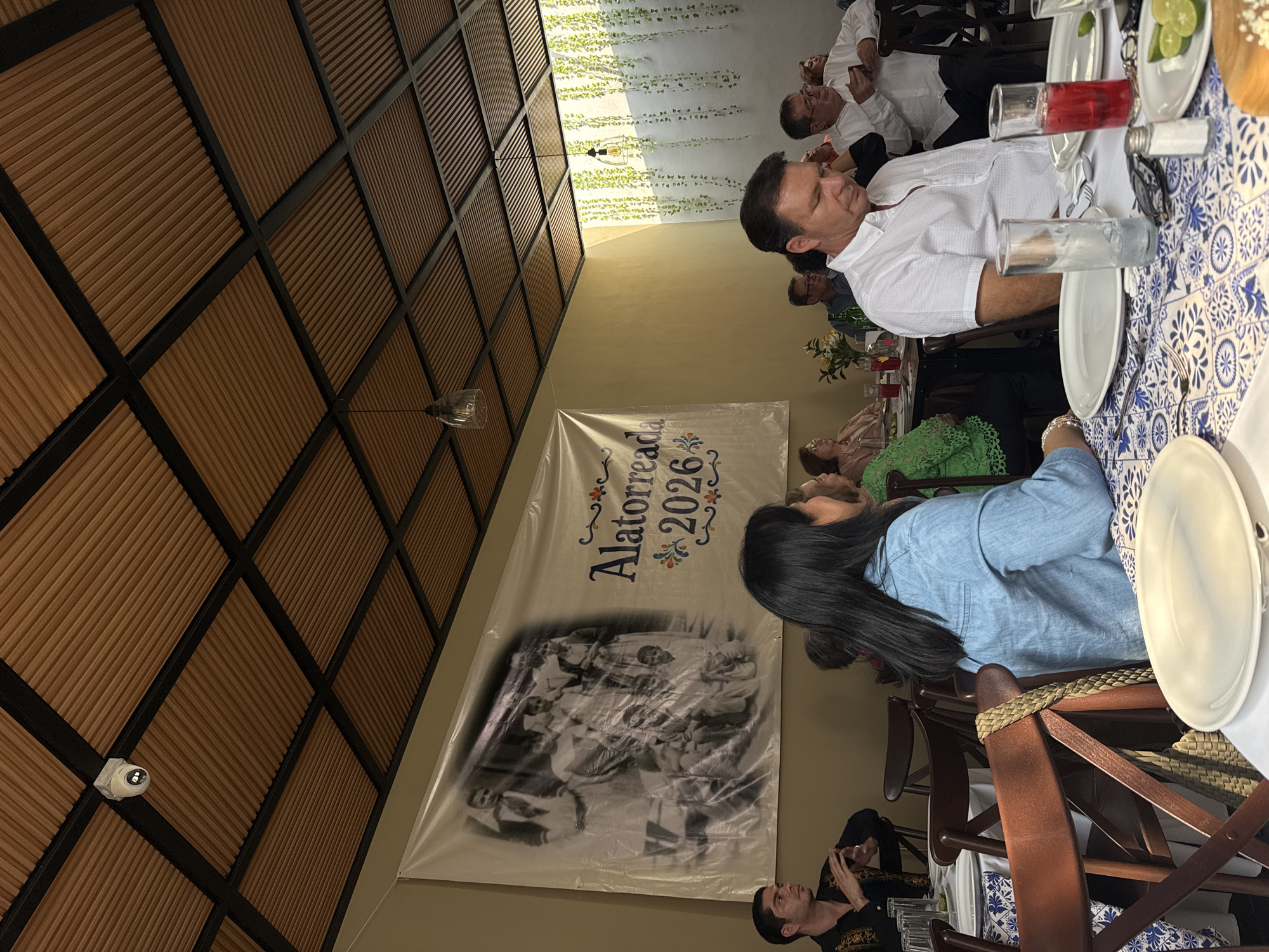 The image shows a group of people gathered for a meal in San Bernardino Tlaxcalancingo, Mexico. A long table with a blue and white patterned tablecloth is set with plates, glasses, and cutlery. Several individuals are seated at the table, including men in white shirts and a woman in a blue shirt. A banner with the text "Alatorre 2026" and a black and white photograph hangs on the wall. A performer in traditional attire is standing near the banner. The location appears to be a restaurant or event space with a decorative ceiling and wall decorations.