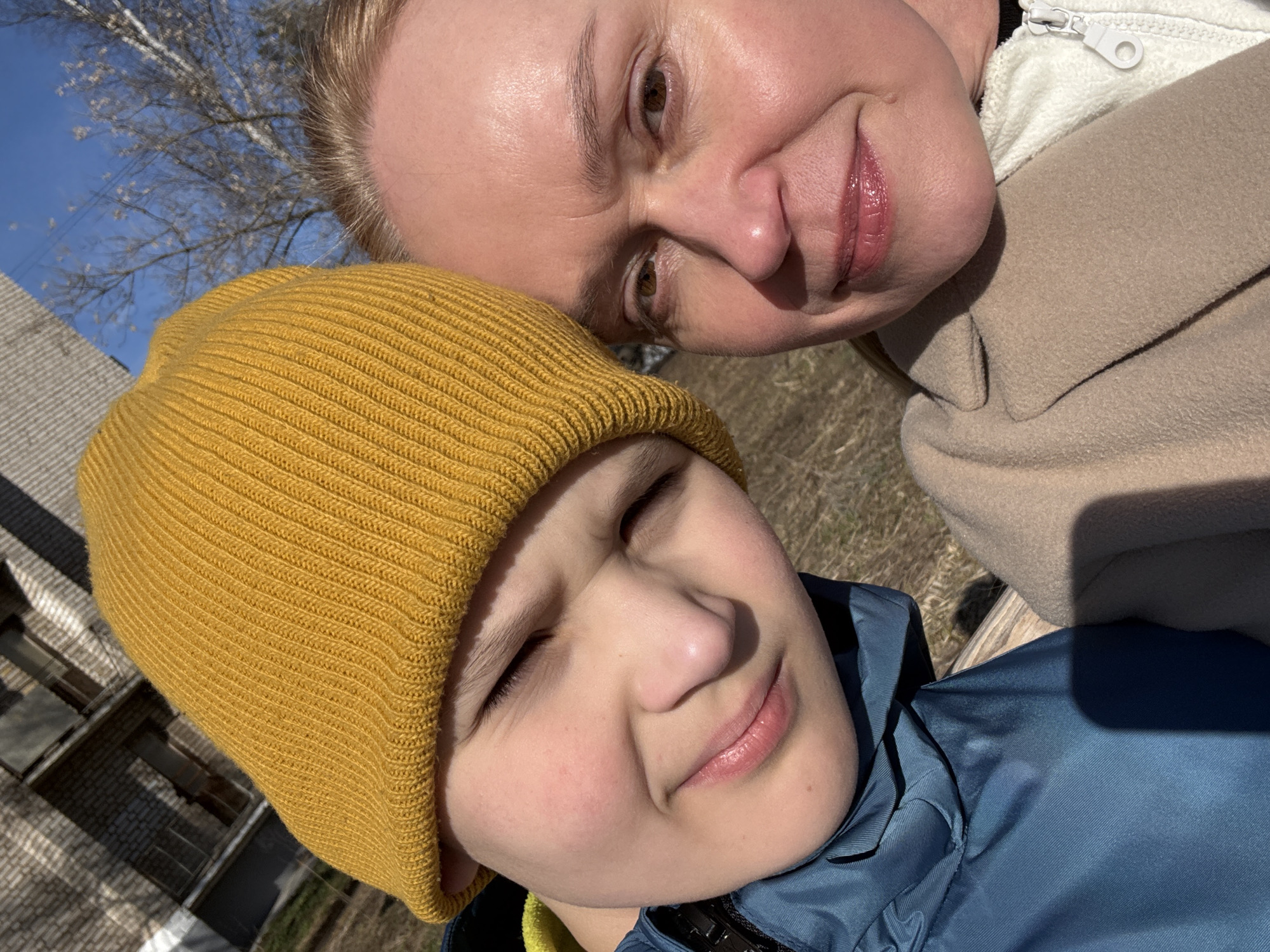 The image is a selfie of a woman and a child in what appears to be Chaikovsky, Russia. The woman is fair-skinned with blonde hair and is wearing a tan coat over a white zippered top. The child is wearing a mustard-yellow knit hat and a blue jacket. The background includes a view of a building, a tree, and a blue sky. The location is likely outside, given the natural light and the outdoor elements in the background.