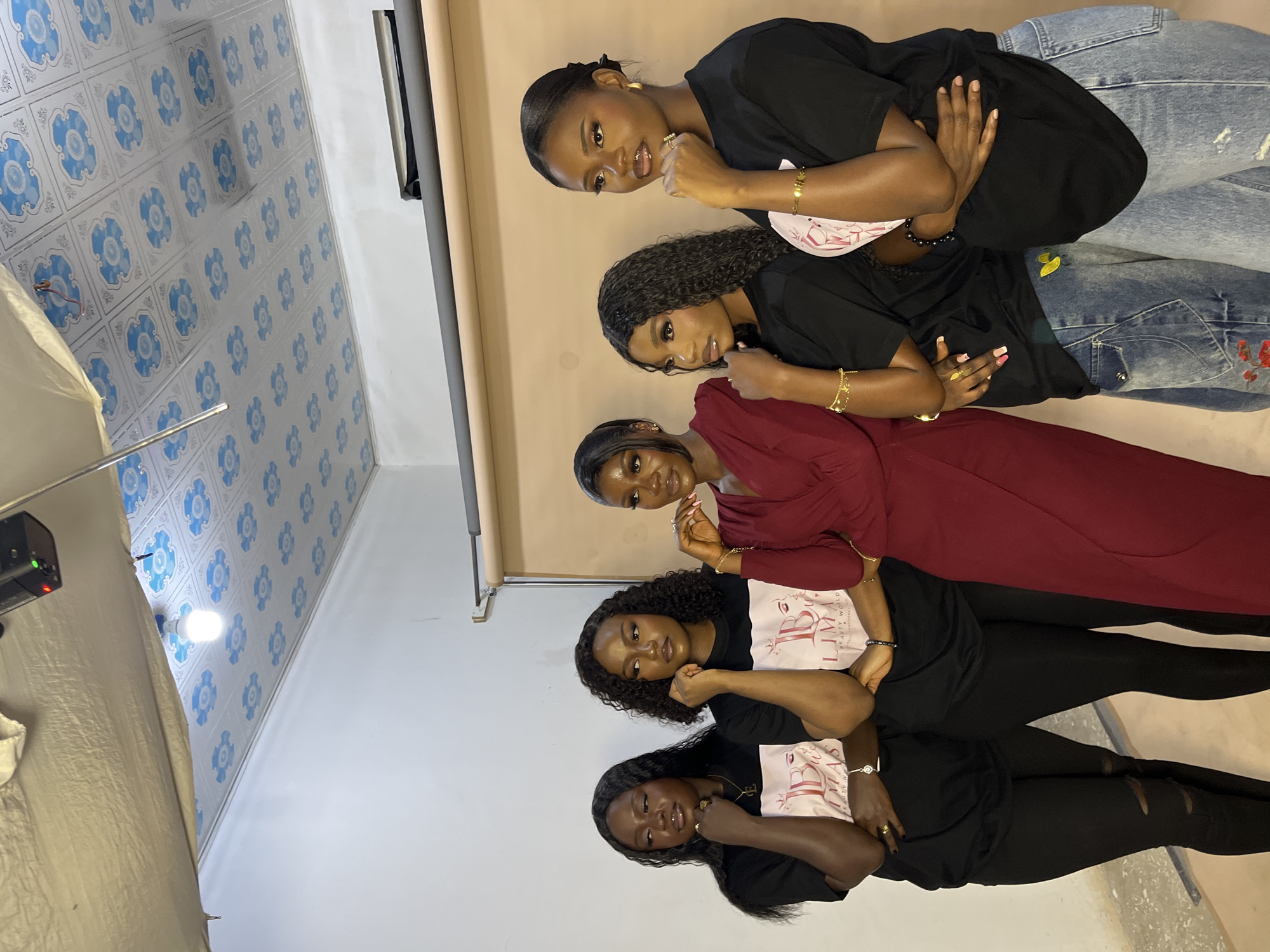 In this media file, five young Black women are posed in a studio setting. They are positioned one behind the other, facing the camera at a slight angle. The background consists of a plain, light-colored backdrop and a section of tiled wall with a blue floral pattern. To the left of the frame, a photography light and a white cloth are visible, suggesting a professional setup.
The women are dressed in casual attire. Three of them wear black t-shirts with a pink logo featuring "LB BEAUTY WORLD" and some stylized text. The woman second from the top is wearing a red dress, and the woman at the very top is wearing a black t-shirt and blue distressed jeans. Several of the women are adorned with jewelry, including bracelets and rings. Their makeup is applied, and they appear to be posing for a photograph, with some resting their chins on their hands and looking towards the camera. The overall mood appears to be poised and confident. The lighting suggests it could be daytime, indoors. The mention of Gura Top, Nigeria, provides a geographical context for the image.