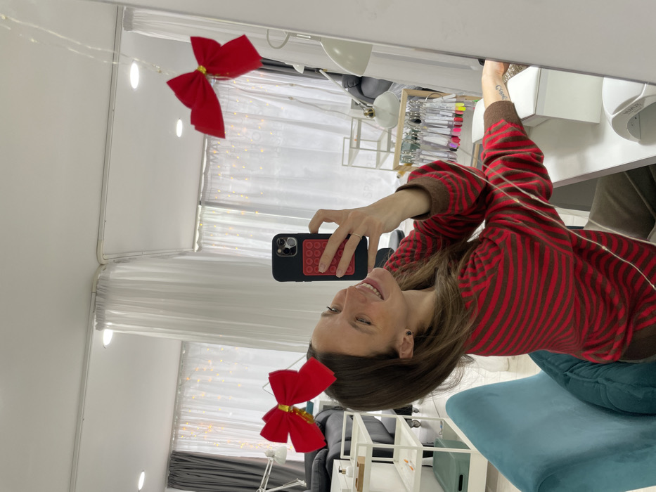 A female subject, appearing to be an adult, is depicted in a mirror selfie within an indoor setting resembling a nail salon or beauty studio. She is smiling, looking at the camera of a black smartphone held in her right hand; the phone case features a red circular "pop-it" style grid. The subject wears a red and brown horizontally striped long-sleeved top and a red headband with two red bows. A silver-colored ring is visible on her left ring finger. The immediate foreground includes a white desk with a white UV nail lamp and a wooden stand displaying multiple nail polish swatch sticks. The subject is seated on a teal upholstered chair. The reflected background features white sheer curtains adorned with fairy lights, a white multi-tiered shelving unit, a dark upholstered armchair, and a white task lamp on a rolling stand. Ceiling lights illuminate the space. The image contains no visual information to confirm a specific location within Budva, Montenegro.