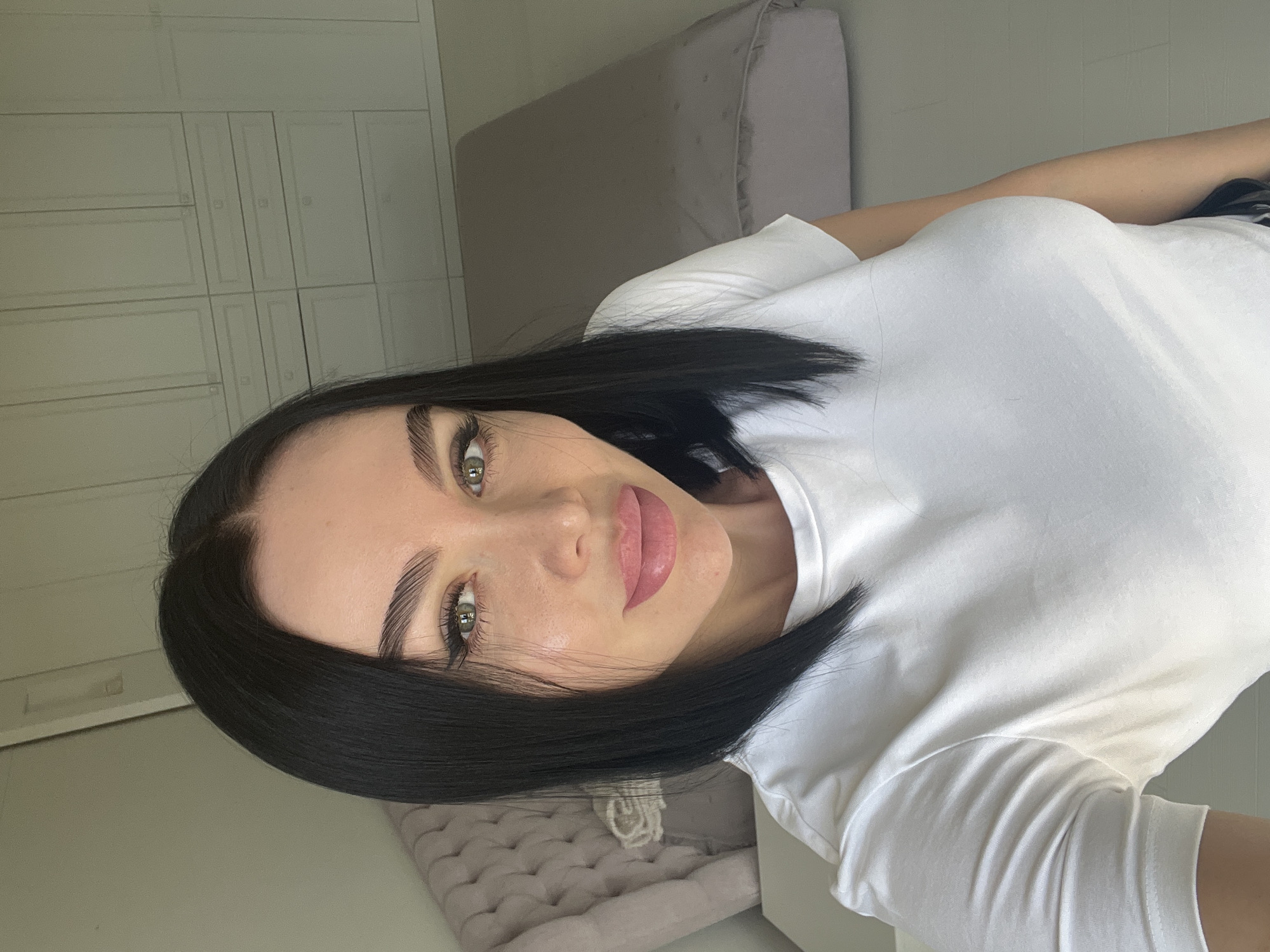 A woman with shoulder-length black hair and green eyes is pictured in a close-up selfie. She is wearing a white t-shirt. Her lips are painted pink, and she has makeup on her eyes, including winged eyeliner. She is looking directly at the camera. The background appears to be a bedroom with a bed and a closet.