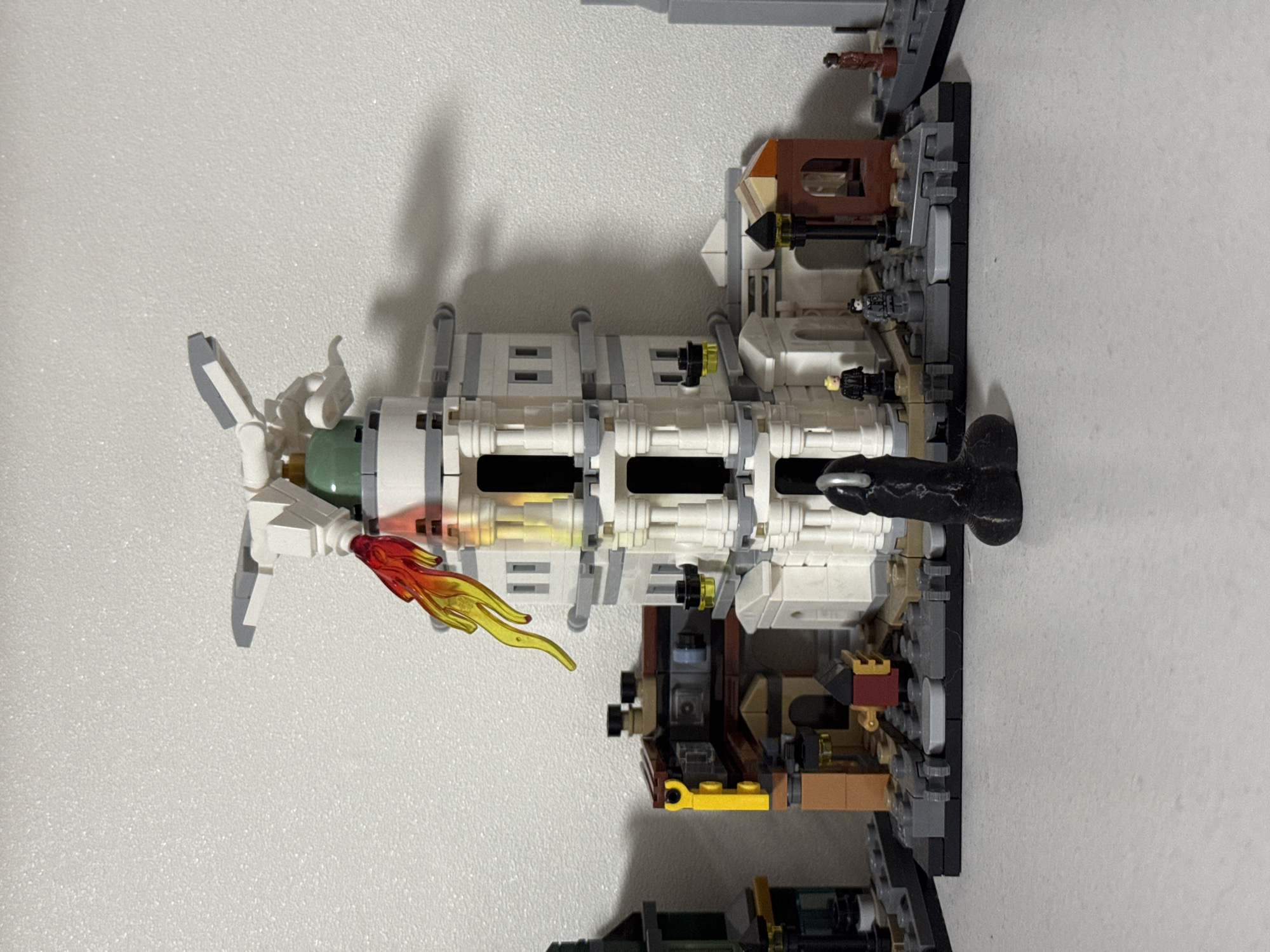 The image shows a LEGO construction of a lighthouse or tower with surrounding buildings. A flame-like red and yellow LEGO piece is attached to the top of the lighthouse, appearing to be emitting from a part of the structure. Several LEGO minifigures are placed around the base of the lighthouse. In the foreground, a black phallic object is positioned on the surface in front of the LEGO creation. The background consists of a plain, textured white wall, suggesting the display is indoors. There is no visible indication of the city of Kyiv, Ukraine.