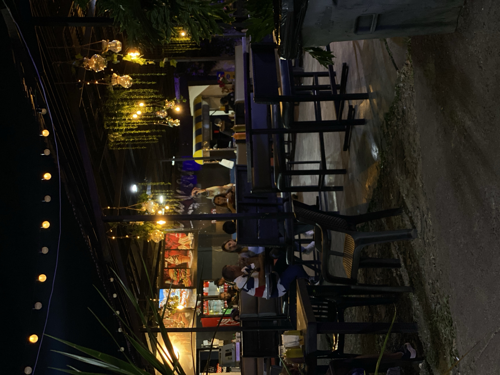 The image depicts an outdoor dining area at night, likely in Tinaquillo, Venezuela. Several people are seated at tables, suggesting a restaurant or cafe setting. The tables and chairs are dark in color. String lights are hung overhead, providing illumination and creating a festive atmosphere. The surrounding environment appears to be a mix of built structures and natural elements, with vegetation visible. The ground is paved or covered with a hard surface.