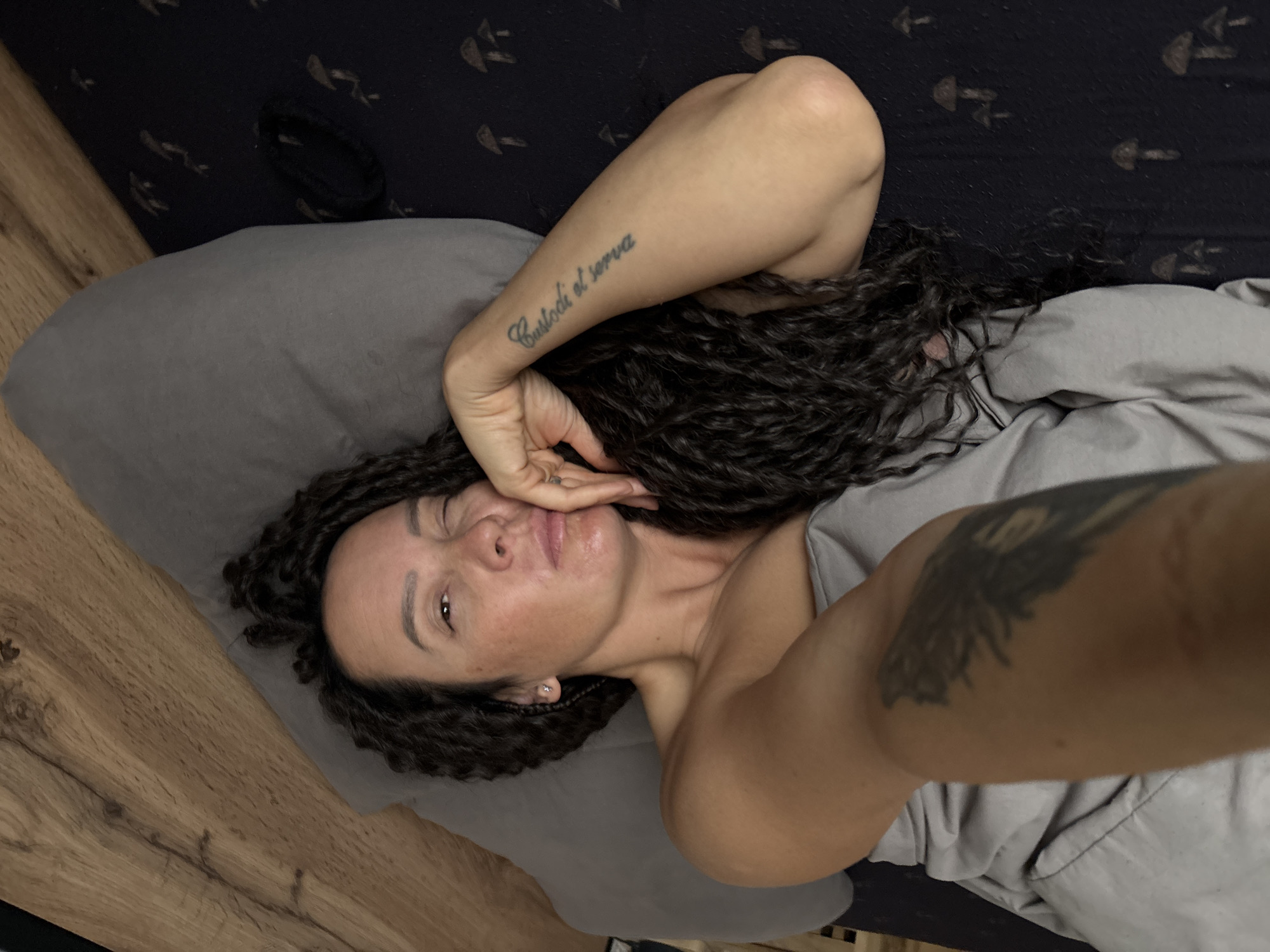 A woman with dark, curly hair and light-toned skin is lying on a bed, looking towards the camera in a selfie-style shot. Her expression is neutral and relaxed, with her right hand gently touching her chin/cheek. She is partially covered by light gray bedding, which also serves as her pillow. A small stud earring is visible in her left ear, and she wears a ring on her right hand.
On her right forearm, there is a tattoo in script that reads "Custodi et serva." Another, larger, dark tattoo is visible on her left shoulder or upper arm, appearing abstract or possibly depicting a creature with wings, though details are indistinct.
The background behind her head features dark bedding with a repeating pattern of light-colored, arrow-like or mushroom-shaped motifs. A black hair tie or scrunchie is also visible on this dark bedding. To the left of the bed, a light brown wooden surface, possibly a floor or bedside table, is visible. The scene depicts an indoor setting, likely a bedroom, in Chaikovsky, Russia.