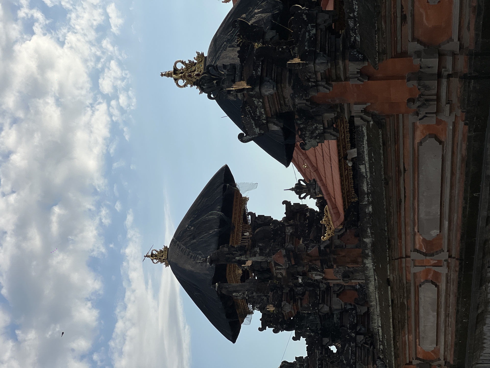 The photograph shows two Balinese pagoda-like structures, likely part of the Taman Ayun Temple in Mengwi, Bali, Indonesia. They are viewed from below, with the sky featuring fluffy white clouds and patches of blue in the background. The structures are predominantly dark in color, adorned with intricate carvings and topped with golden ornaments. The structures appear to have multi-tiered roofs. Parts of the building materials are reddish-orange. The perspective makes the structures appear imposing against the open sky.