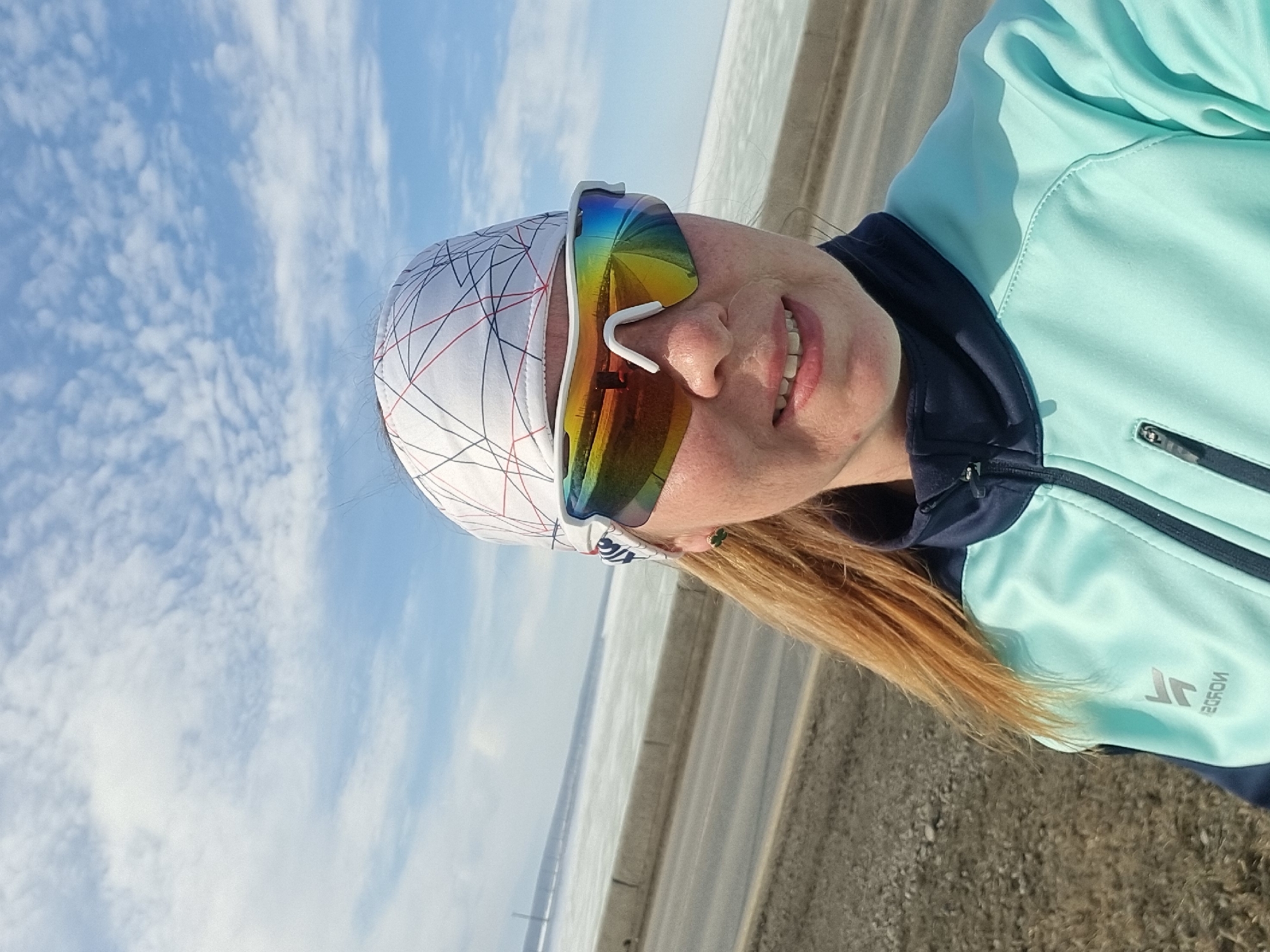 The image displays a woman in a selfie photograph taken outdoors in Chaikovsky, Russia. The subject, a woman with blonde hair, fair complexion, and a slight smile showing her teeth, occupies the right half of the frame. She wears a white headwrap patterned with black and red intersecting lines, white-framed sports sunglasses featuring rainbow-mirrored lenses, and a light aqua-teal full-zip athletic jacket with a dark blue collar and zipper accents. A logo is visible on the left chest of her jacket. A small green earring is present on her visible ear. The background features a paved road running from the mid-left to the center, bordered by a low concrete barrier. Beyond the barrier, a wide body of water, partially covered in white ice or snow, extends to the horizon. The sky above is light blue with scattered white clouds. A faint, distant structure is discernible on the horizon line across the water. The immediate foreground to the left of the woman consists of dry, brown earth.