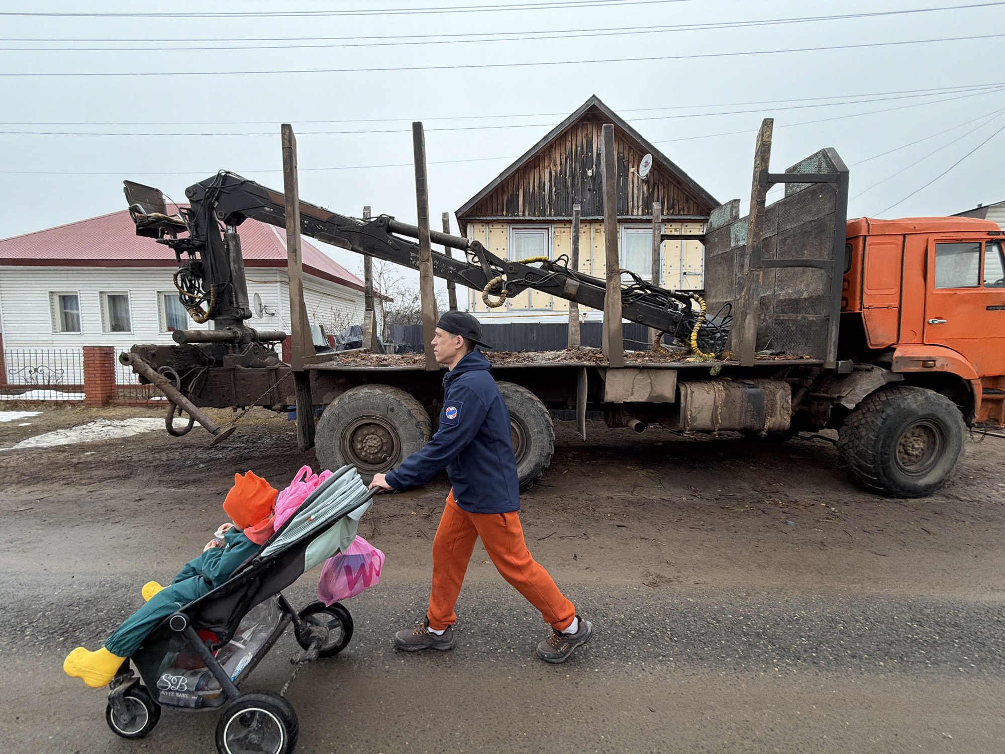 A person wearing a dark blue jacket, rust-colored pants, and a black cap is pushing a stroller down a street. The stroller contains a small child wearing an orange hat and yellow boots. Two shopping bags are hanging from the stroller. To the right is a large orange truck with a crane and logging equipment mounted on the back. The truck appears to be parked on the side of the road. Behind the truck are two houses, one white with a red roof and the other made of unfinished wood. The street is wet and muddy, indicating recent precipitation. The photograph was taken in the city of Chaikovsky, Russia. The overall impression is of a rural or semi-rural setting.