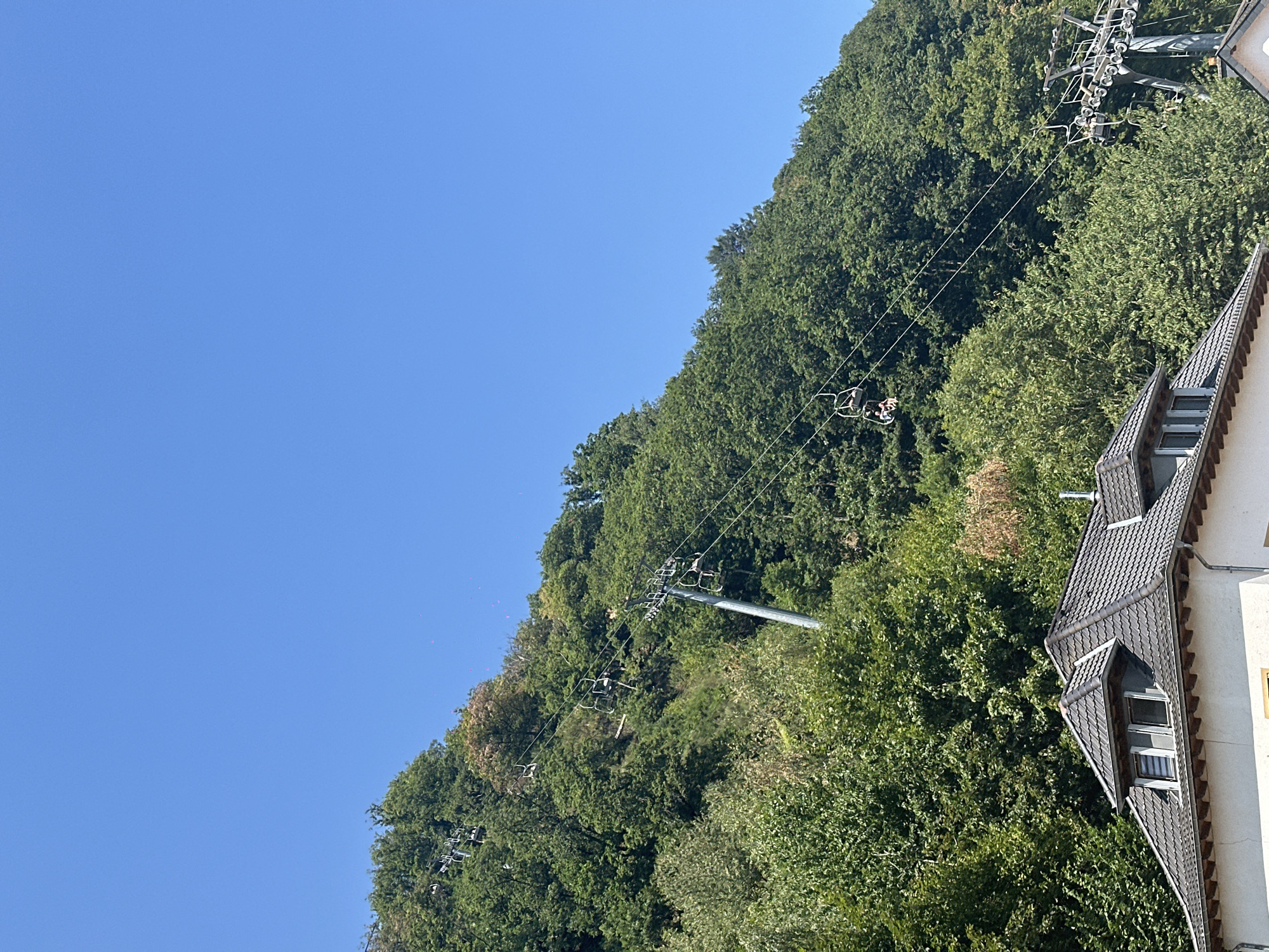 The image shows a ski lift ascending a tree-covered hill under a clear blue sky. The ski lift consists of a series of chairs suspended from a cable, with several chairs occupied by people. In the foreground, the roof of a building is visible, suggesting a residential or commercial area at the base of the hill. The lush green foliage indicates a summer or spring season. This scene is characteristic of the surroundings of Vianden Castle in Vianden, Luxembourg, where a chairlift operates, providing access to scenic viewpoints and hiking trails. The overall impression is one of a recreational area offering panoramic views of the landscape.