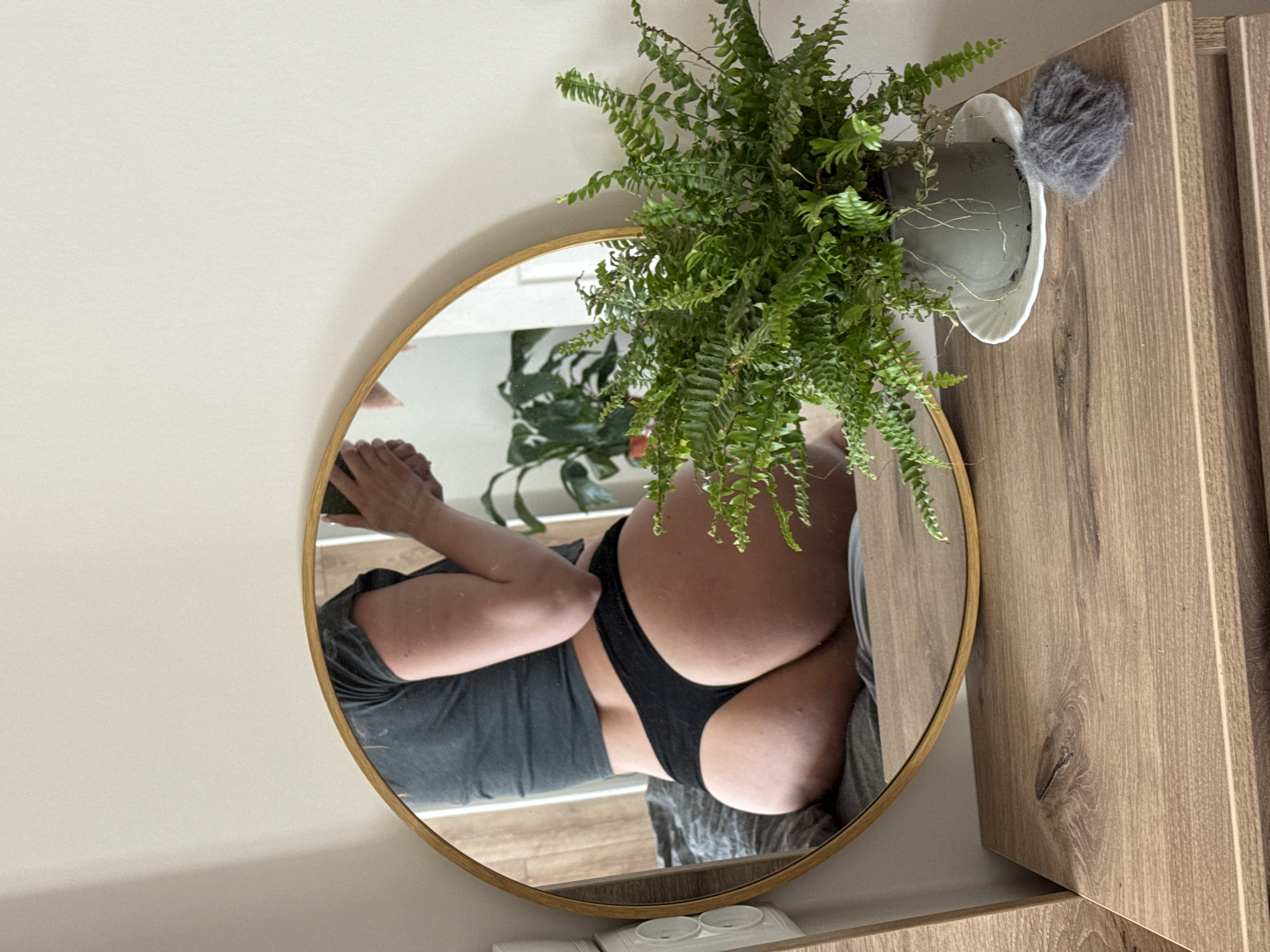 A low-angle, medium shot captures a reflection in a round, gold-framed mirror of a person's lower body. The person is wearing a dark grey crop top and black thong underwear. They are sitting on a bed with grey bedding, and their back is to the viewer. The mirror is positioned on a wooden dresser. To the right of the mirror, a potted fern with vibrant green leaves sits on the dresser. A small clump of grey fluffy material is also on the dresser, near the fern. The background consists of a plain, light-colored wall. The lighting suggests an indoor setting, possibly a bedroom. There is no indication of the city or location.
