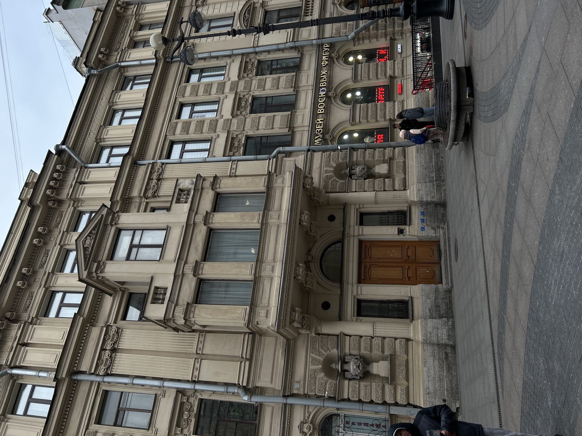 The image shows a beige building located in Saint Petersburg, Russia. The building is multi-storied and has architectural details, including window frames, moldings, and drainpipes. The entrance features a large wooden door under a curved arch. Above the entrance, a sign in Russian reads "МУЗЕЙ ВОСКОВЫХ ФИГУР," which translates to "Wax Figures Museum." A group of people is visible on a bench in front of the building. The street is paved with gray cobblestones. A man lying down is visible near the bottom of the image. The sky is overcast.
