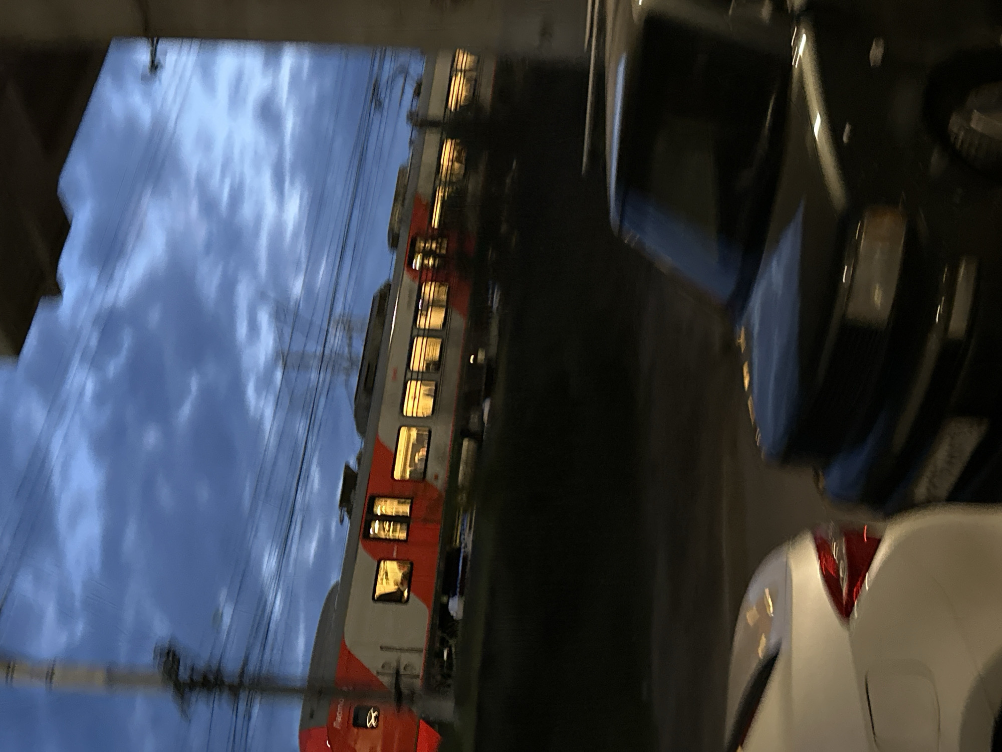 The image captures a twilight or early evening scene featuring a passenger train and vehicles. The photograph is oriented vertically, but when viewed horizontally, it depicts a long electric train with illuminated windows, running from left to right across the middle of the frame. Above the train, a cloudy, blue-grey sky characteristic of dusk or dawn dominates the background.
The train itself is a modern, multiple-unit passenger type, distinguished by its white, grey, and red livery. The numerous windows along its side glow warmly from interior lights, and indistinct figures of passengers can be seen within some carriages. Faint text "Ласточка" (Lastochka), a common Russian electric train model, is visible on the lower red section of the train. Overhead power lines (catenary) necessary for electric trains are prominent above the tracks.
To the right of the train, two cars are partially visible. Closer to the top right is a dark-colored SUV or truck. Below it, towards the bottom right, is the rear section of a white car, possibly a sedan, with its red taillight illuminated. Utility poles and some dark, blurry vegetation line the railway environment. The overall mood is calm and atmospheric, with the artificial lights of the train and cars contrasting with the fading natural light of the cloudy sky. The "Ласточка" train strongly suggests the scene's location in Russia, aligning with the provided context of Sochi.