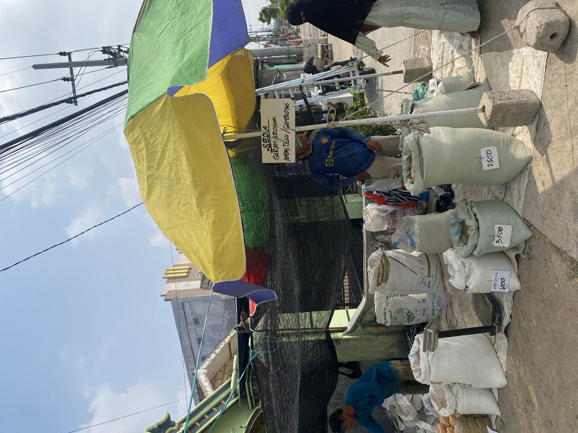 The image depicts an outdoor market stall in Sempol, Indonesia. A man in a blue t-shirt is sorting through white sacks of goods. A woman in a light-colored dress and headscarf is interacting with the stall, pointing at a sack. Several large, light-colored sacks are arranged on the ground, some with handwritten labels indicating prices. A scale is placed between two of the sacks. Above the stall, a colorful umbrella shades the area, and a sign reads "SEDIA GARAM EROSOK AMPASTELO/GAMBLONG." The background shows buildings, power lines, and a street, suggesting an urban or semi-urban environment. The overall scene captures a moment of commerce in a local Indonesian marketplace.