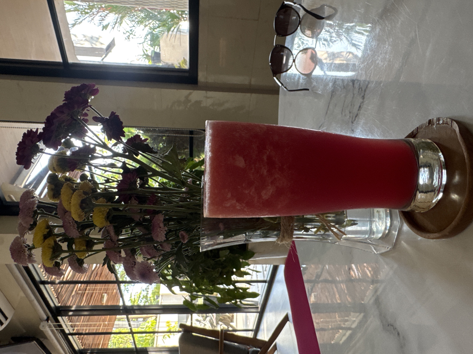 The image depicts a table setting, likely at a cafe or restaurant in Kuta, Indonesia. Dominating the foreground are a tall glass of a red beverage, probably juice, and a vase filled with purple and yellow flowers. Behind them is a window showing green vegetation. A pair of sunglasses rests on the table’s surface beside the drink.