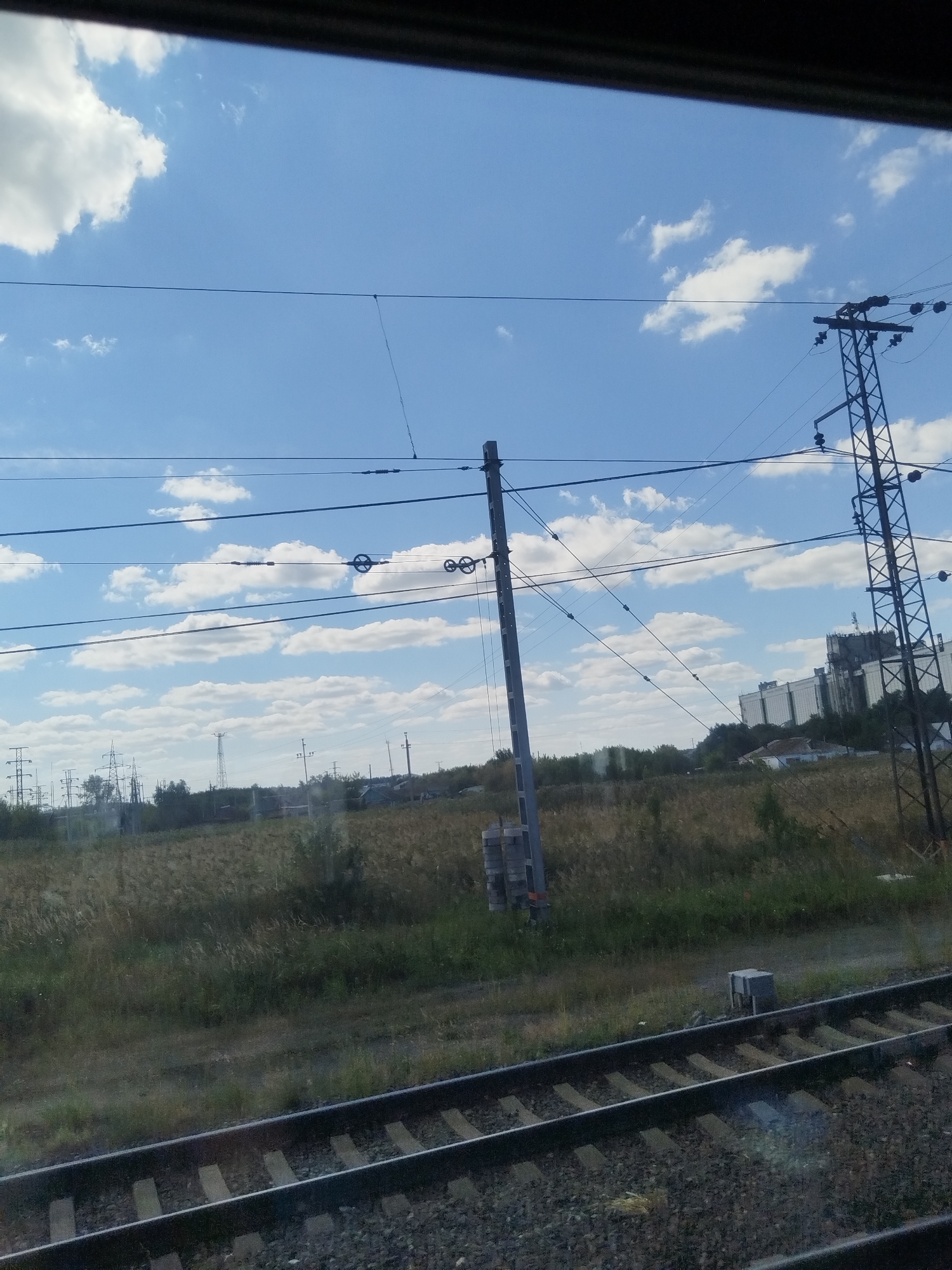 The image depicts a view from a train traveling through Moskalenki, Russia, on a clear, cloudy day. In the foreground, train tracks are visible, with ballast and wooden sleepers. Beyond the tracks, a field of dry grass and sparse green vegetation stretches out. Several utility poles with wires are scattered throughout the landscape, along with some distant buildings and structures, possibly part of the town. In the far background, a large industrial building complex is discernible. The sky is a vibrant blue, dotted with scattered white clouds. The overall scene suggests an industrial or semi-rural area on the outskirts of Moskalenki. There are no people visible in the image.