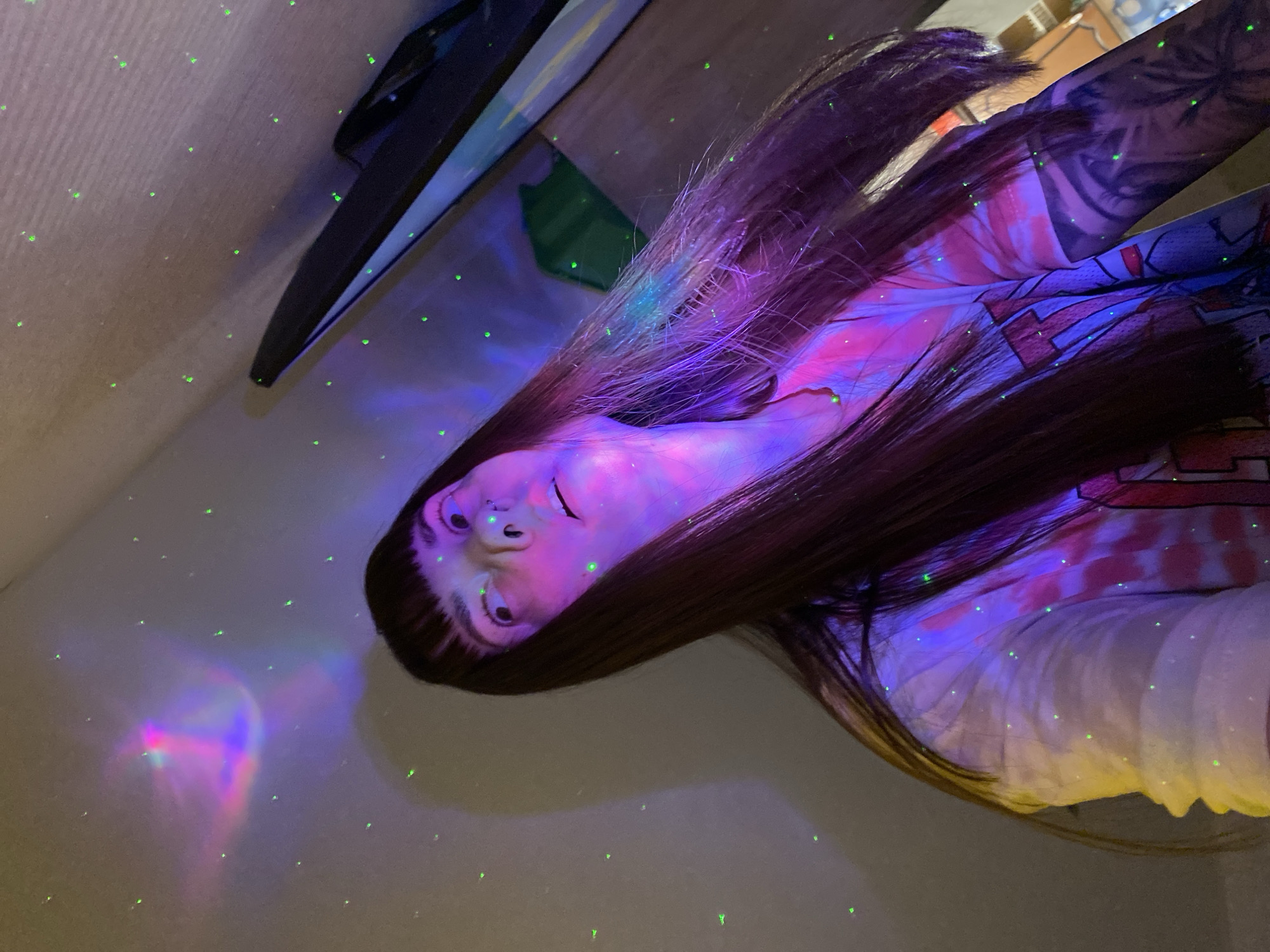 A person with long, dark hair is lying on their side, their face illuminated by a purple and blue light. The light also shines on their shirt, which has a tie-dye pattern and a graphic design. The person has tattoos on their arm. Small green dots of light are scattered across the wall and ceiling. A television and a green box are visible in the background. The location of the image is unknown and cannot be determined from the available information.