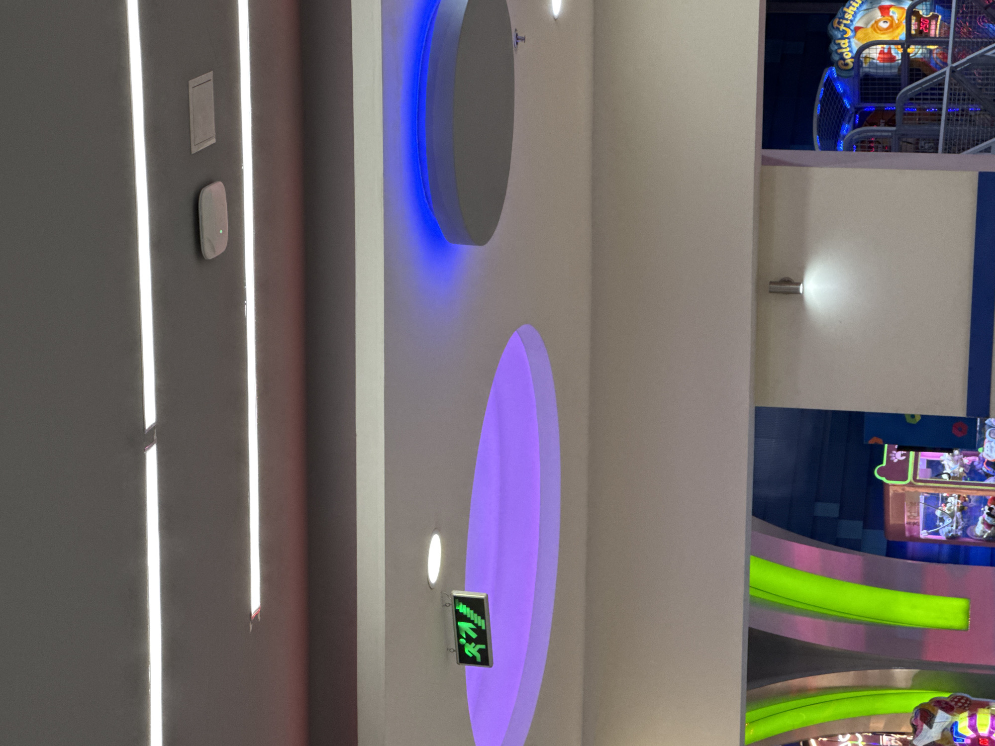 This image captures a vibrant and modern indoor setting, likely an entertainment center or arcade, characterized by diverse artificial lighting. The scene is devoid of people.
On the left side, a light gray wall features several prominent fixtures. Two bright, vertical LED strips run parallel, illuminating the surface. Between them, a small rectangular panel is mounted above a smaller, oval-shaped device with a glowing green indicator light. Further to the right, two large, circular disc-like fixtures protrude from the wall. The upper disc is backlit with an intense blue glow, while the lower one emits a soft purple light. Below the purple-lit disc, a green exit sign is visible, displaying a running person pictogram and an arrow pointing downwards, indicating an emergency exit.
The right side of the image reveals glimpses of a colorful arcade or play area. In the top right corner, part of an arcade game is visible, featuring a golden sign that reads "Gold Fishin'." Blue lights adorn this structure, and a digital display shows "250". Below this, a small, cylindrical silver light fixture projects a bright white light onto the wall. The bottom right section showcases bright green, curved, illuminated structures, possibly part of seating or decorative elements, along with other partially visible game components.
The overall ambiance is energetic and playful, created by the contrasting and vibrant artificial lights. The setting suggests a place designed for recreation and leisure, possibly during the evening hours. The visible text includes "Gold Fishin'," "250," and the international exit pictogram.