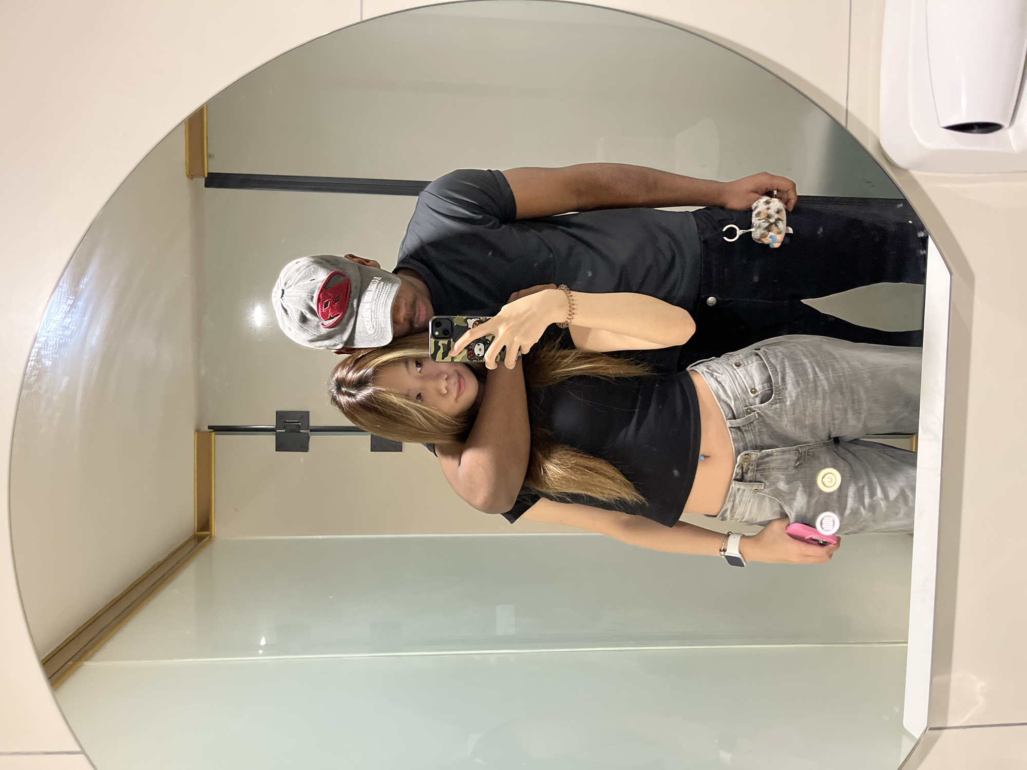 A mirror selfie captures a man and a woman posing together in what appears to be a public restroom or changing area in Yuzhong District, China. The large, round mirror reflects the subjects and parts of the surrounding environment.
The man, on the left, wears a grey baseball cap with a red design and partially visible white text that reads "PROHIBITED AREAS." He is dressed in a black t-shirt and dark pants, with a small, spotted, fluffy keychain attached to his waist. His right arm is wrapped around the woman.
The woman, on the right, has long, light brown hair. She is wearing a black crop top that reveals a belly button piercing, and light grey jeans. On her left wrist, she wears a white smartwatch. She is holding a mobile phone in her left hand, which has a camouflage-patterned case featuring a Hello Kitty design. She also holds a pink object, possibly another phone or a wallet, in her right hand. Both individuals are looking towards the camera within the phone held by the woman, suggesting she is taking the photo.
The background reflected in the mirror consists of light-colored walls, horizontal dark bars or rails, and a white, wall-mounted hand dryer or soap dispenser in the top right. A golden-colored trim is visible along the bottom edge of the reflected wall.