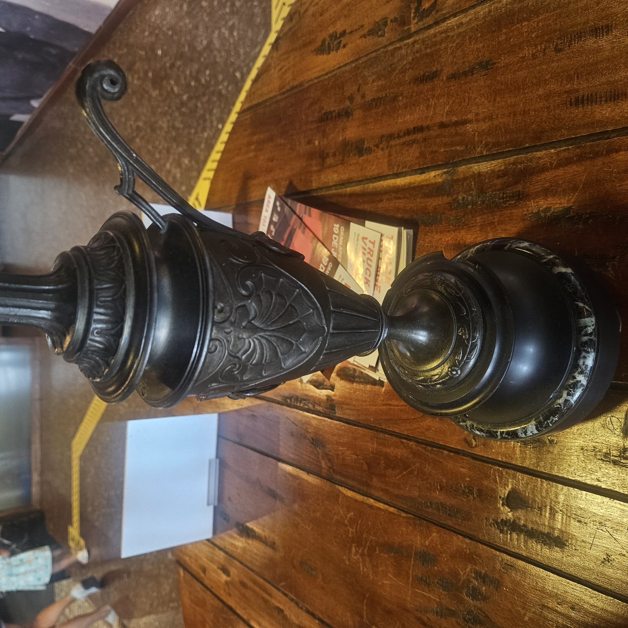 The image shows a close-up of an ornate dark metal lamp on a wooden table. The lamp has decorative carvings and a curled handle on its side. In the background, out of focus, there are glimpses of people, a yellow line on the floor, and a white rectangular object. A stack of papers with some text visible, including the word "TRUCK", is placed beneath the lamp. The lighting suggests an indoor setting with warm tones. The scene is devoid of any specific landmarks or elements that would indicate a location in Almadrones, Spain.