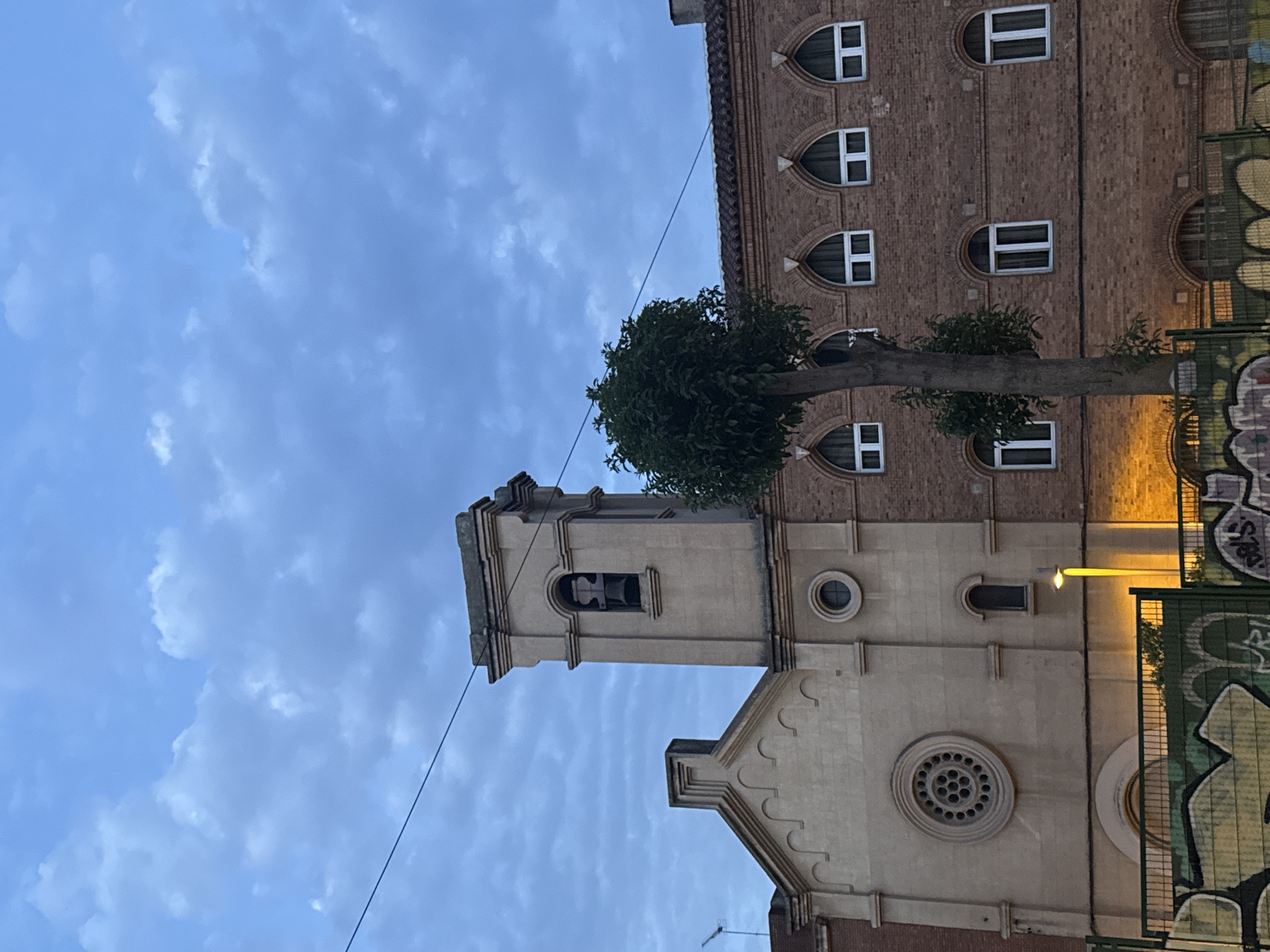 The image shows the façade of a building in Barcelona, Spain, with a bell tower and a brick extension. The sky is overcast with clouds. In the foreground, there is a fence with graffiti. A tree stands in front of the brick extension. The building appears to be a church or a similar religious institution. The architecture suggests it is an older building. The streetlamp indicates it is either dusk or early morning. There are no people visible in the image.