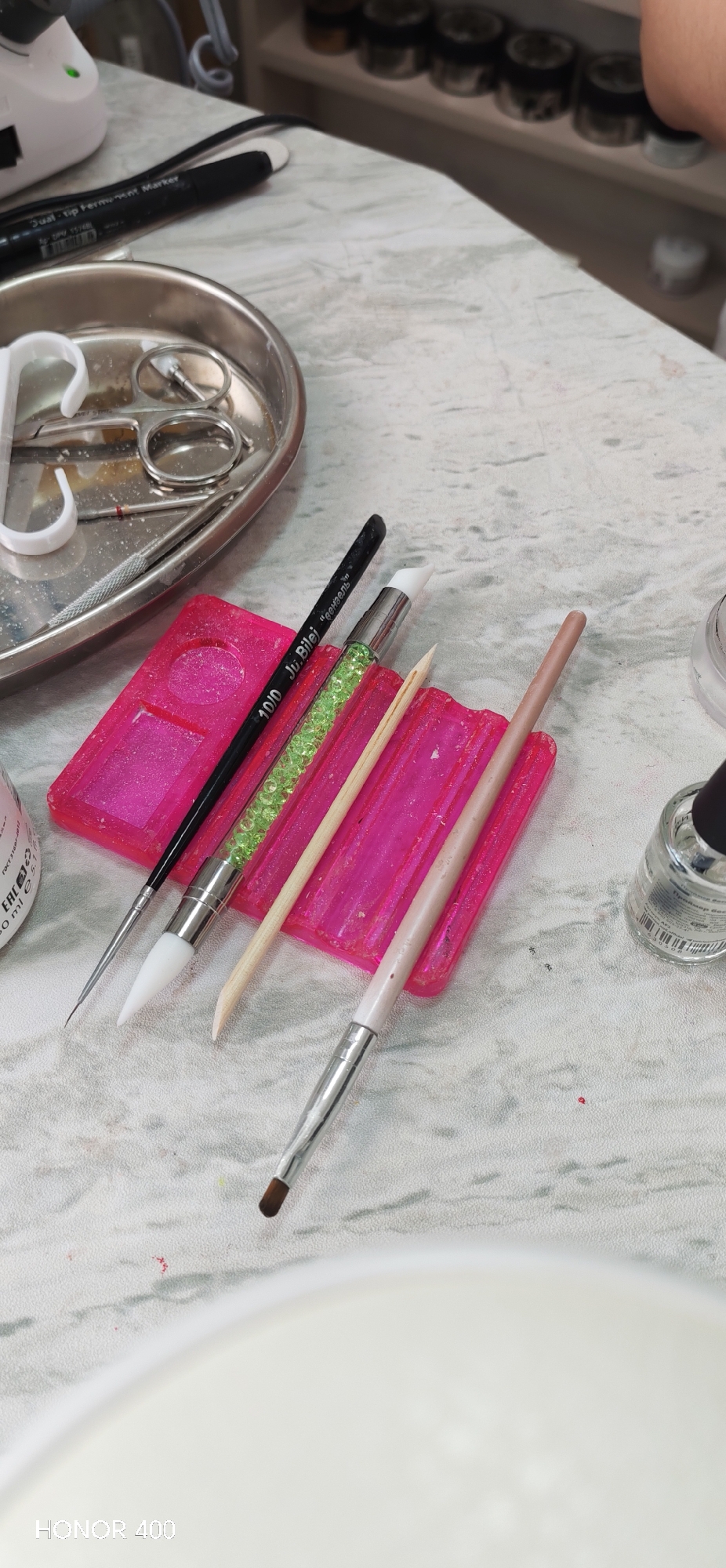 The image shows a workspace with manicure tools. A small stainless steel tray contains scissors, a metal cuticle pusher, and other metal implements. A pink plastic holder holds a thin black nail art brush labeled "10/0 Ju. Bilej", a green-jeweled brush with a white silicone tip, a wooden cuticle stick, and a wide silver-handled nail brush. There is also a bottle of clear nail product nearby. A black marker is partially visible above the tray. Background shelves hold dark jars. "HONOR 400" is printed in the lower left corner. The location is not discernible in the provided image.