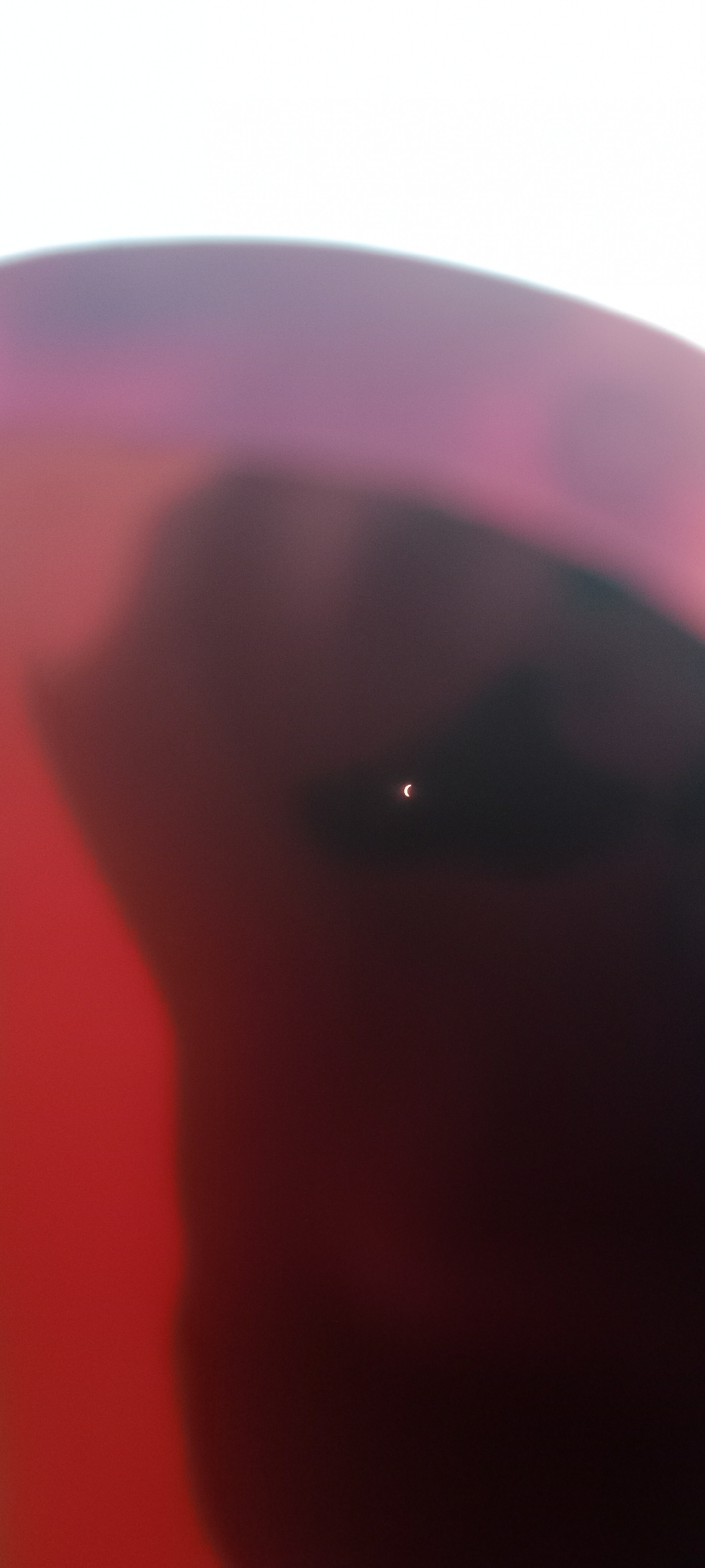 The image shows a sliver of a solar eclipse, appearing as a crescent-shaped light, set against a dark, out-of-focus background with a reddish hue. The top of the image is bright white, suggesting the sun or a very bright light source. There are no discernible people, objects, or actions occurring in the scene. The location in the city of Adai, Spain, is not indicated by any visual cues in the image.