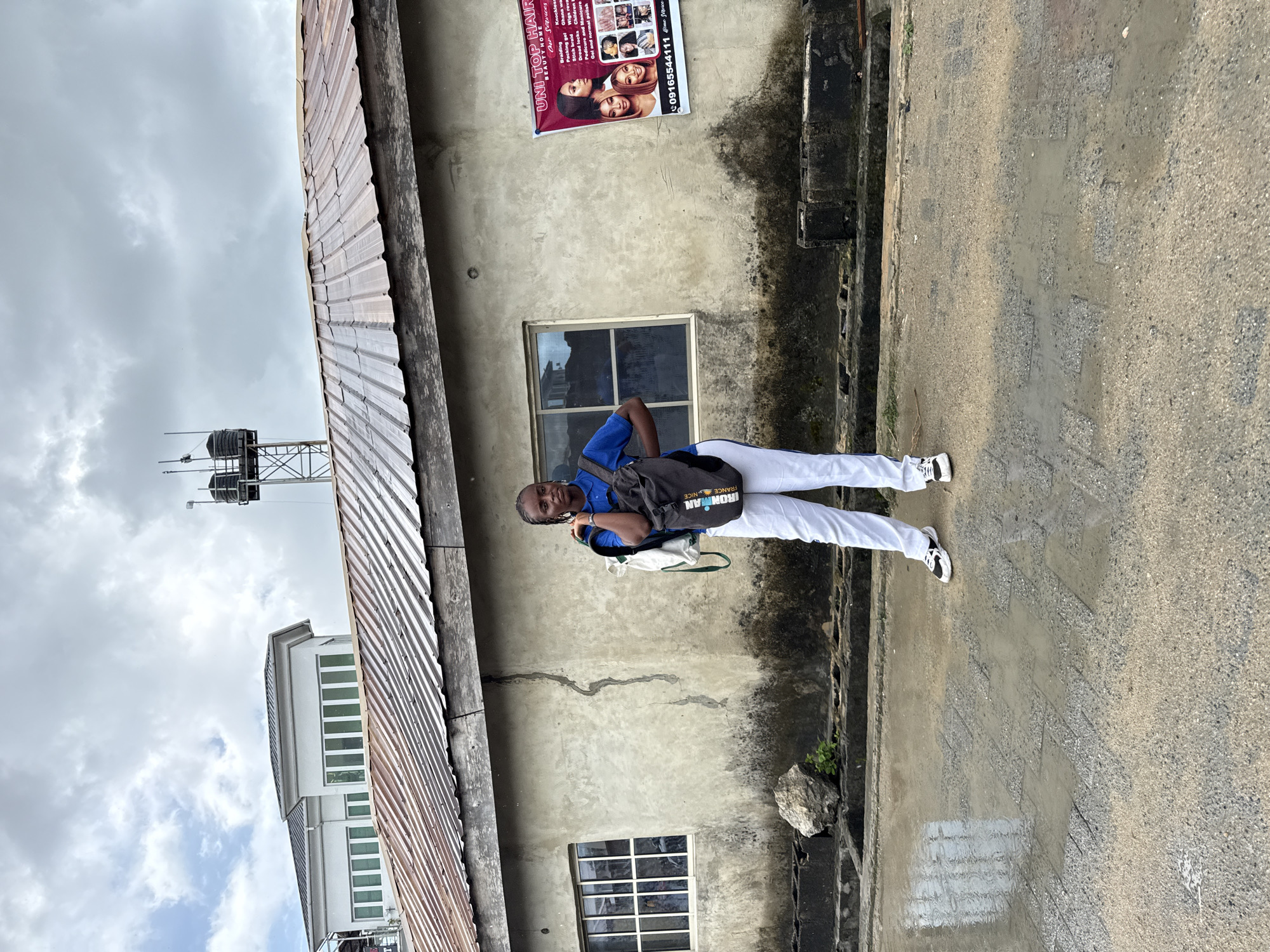 A full-body shot captures a person dressed in white pants and a blue shirt, standing outdoors against a building with a weathered, gray façade. The building features a window, a crack running down its length, and a poster advertising hair services. To the left of the building, a corrugated metal roof is partially visible, with a water tower structure in the background. The person wears a backpack labeled "IRONMAN FRANCE NICE." The ground appears to be a concrete sidewalk or pavement. The scene is possibly located in Ado, Nigeria, given the phone number on the poster matches a Nigerian code. The sky above is overcast with gray clouds.
