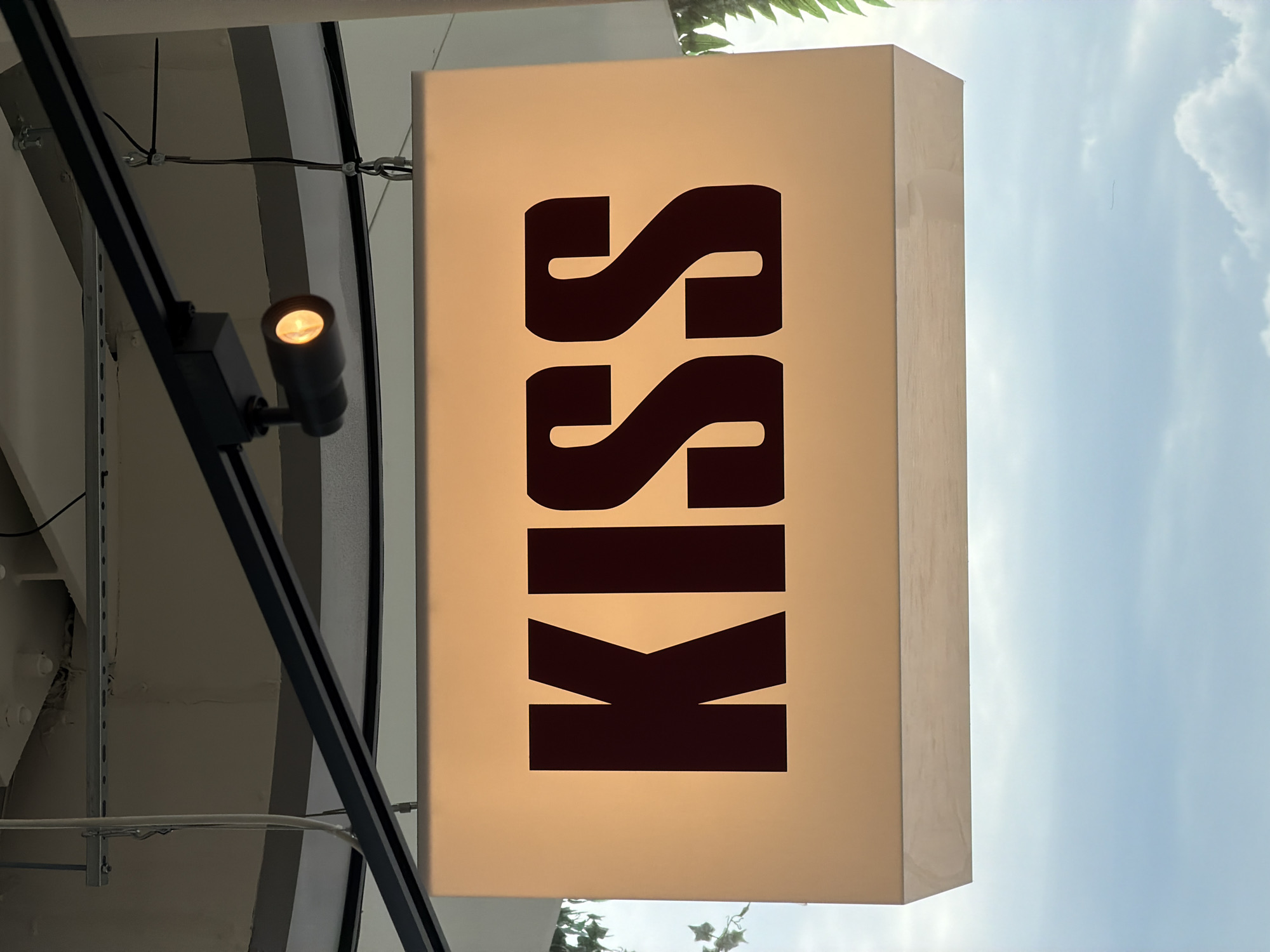 A close-up, medium shot captures a brightly lit, rectangular sign displaying the word "KISS" in large, dark, block letters. The sign is illuminated from behind, casting a warm, orange glow. Above the sign, a black track light is mounted on a black metal beam, directing a single light downwards. The background is a soft, out-of-focus blur of the sky with clouds. The environment appears to be outdoors, possibly a rooftop or an elevated walkway, given the view of the sky and the hanging structure of the sign. There are no people visible in the image. The sign is centrally located in the frame, and the lettering is clearly legible. The image is sharp and well-lit.