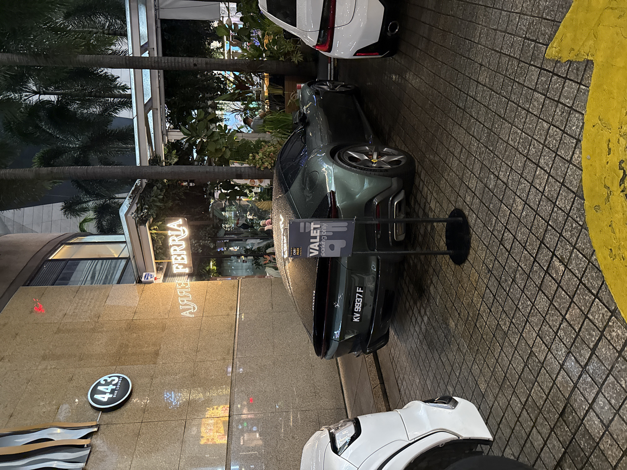 The image shows a valet parking area in front of the building housing the "443 Bar Lounge" and "Amaris Ferria" in Kuala Lumpur, Malaysia. A dark-colored car, displaying the license plate "KV 9937 F", is parked with a valet parking sign nearby. The environment includes palm trees, building structures, and streetlights, suggesting an urban setting. The weather appears to be wet. Additional cars are parked behind and in front of the dark-colored car.