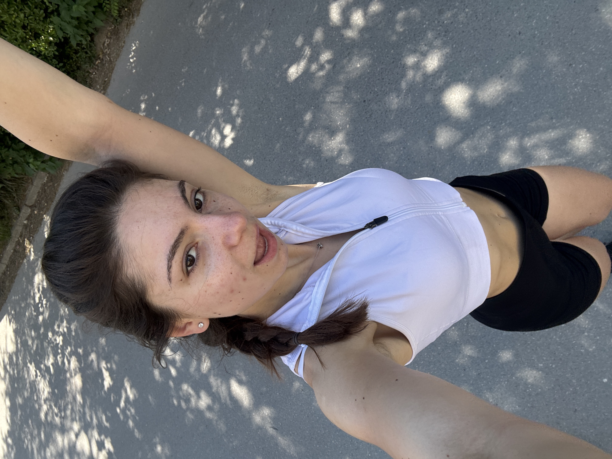 A young woman is seen in a self-taken, slightly distorted overhead shot. The woman has dark hair, light skin with freckles, and is wearing a white athletic top and black shorts. She has a braid in her hair and is wearing an earring. She has her arms raised overhead. The background is a grey paved surface with dappled sunlight, suggesting the presence of trees. The location appears to be an outdoor area, likely a park or walkway in Varna, Bulgaria.
