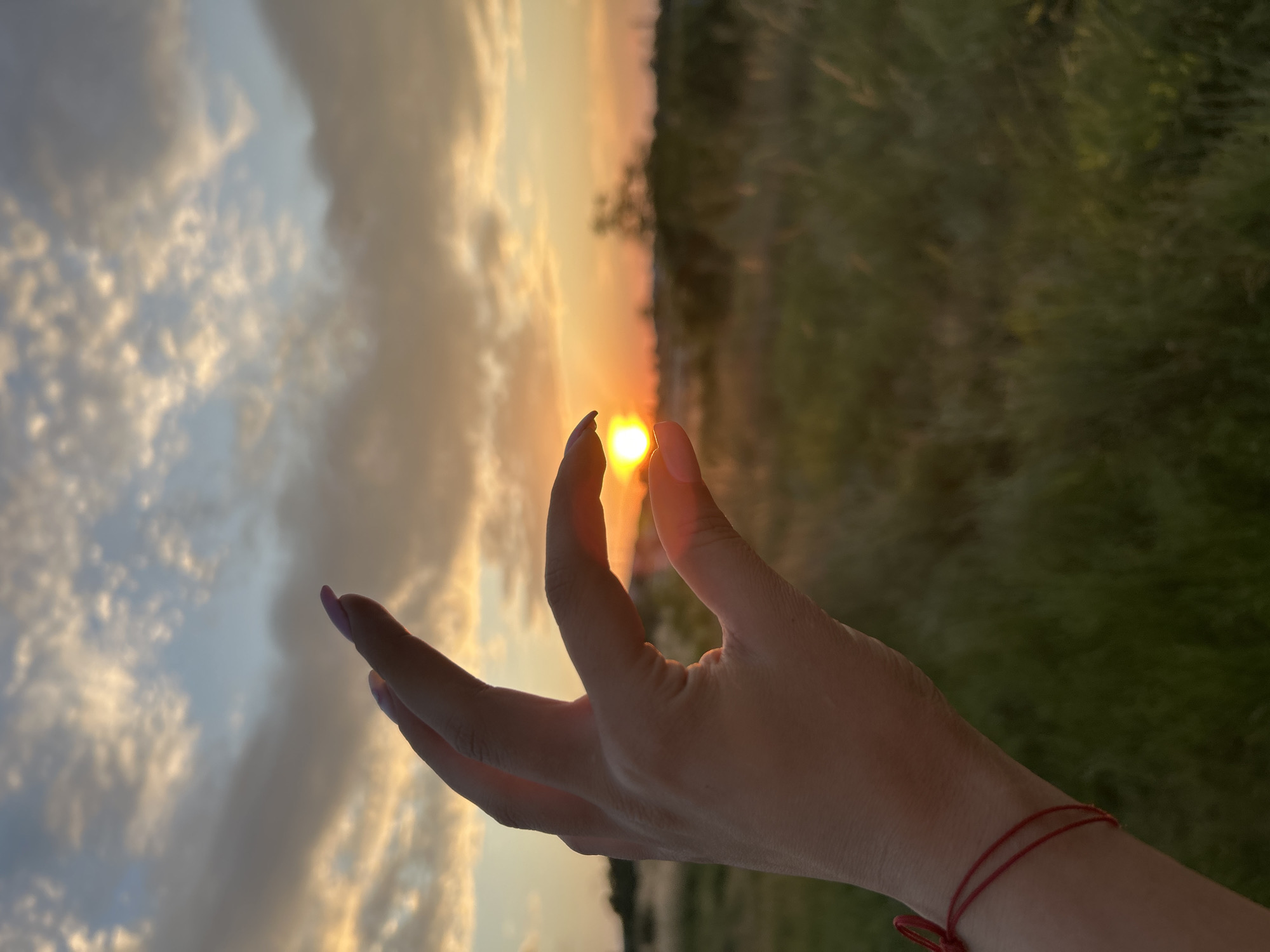 A hand is held up to the sky, with the thumb and index finger forming a circle around the setting sun. The sun is a bright, warm orb, casting a golden glow on the surrounding clouds. The clouds are wispy and varied in color, ranging from pale blue and gray to vibrant orange and yellow, indicating it's either sunrise or sunset. The background shows a blur of green, likely a field or trees, suggesting an outdoor setting. The hand has manicured fingernails, painted a light shade of pink. A thin red string bracelet is tied around the wrist. The overall impression is one of peace and tranquility, with a sense of capturing a moment of natural beauty. The image appears to be taken at a low angle, emphasizing the sky and the hand.