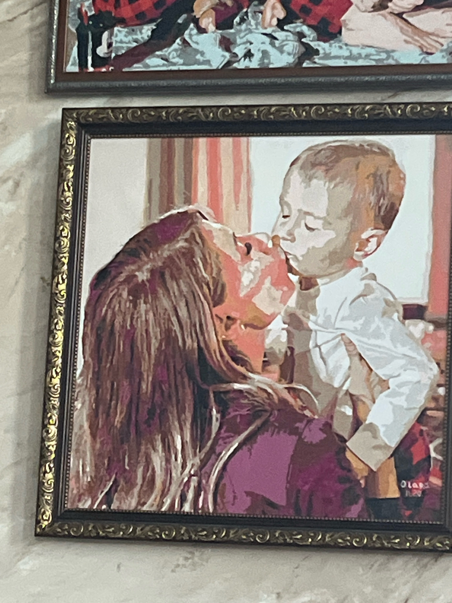 The media file shows two framed artworks. The primary artwork in the foreground is a portrait of a woman and a young child. The woman, with long, dark hair, is leaning in to kiss the child, who is looking upwards with closed eyes. The child is wearing a white shirt. The background of this portrait features vertical stripes in shades of pink and white. The artwork is framed by an ornate, dark frame with gold accents.
Above this portrait is a second, partially visible artwork. This artwork appears to be a more abstract depiction, featuring red and black plaid patterns, and lighter colored shapes. It is also framed, with a darker frame.
The overall scene suggests a home or gallery setting, with a neutral-colored wall as the backdrop. The style of the paintings appears to be illustrative, possibly paint-by-number or a similar artistic technique, given the distinct blocks of color.
There is some faint text visible in the lower right corner of the foreground artwork, possibly a signature or date, but it is largely illegible. No specific time of day, weather, or explicit location cues are evident within the artwork itself. The interaction between the mother and child conveys a sense of affection and tenderness.