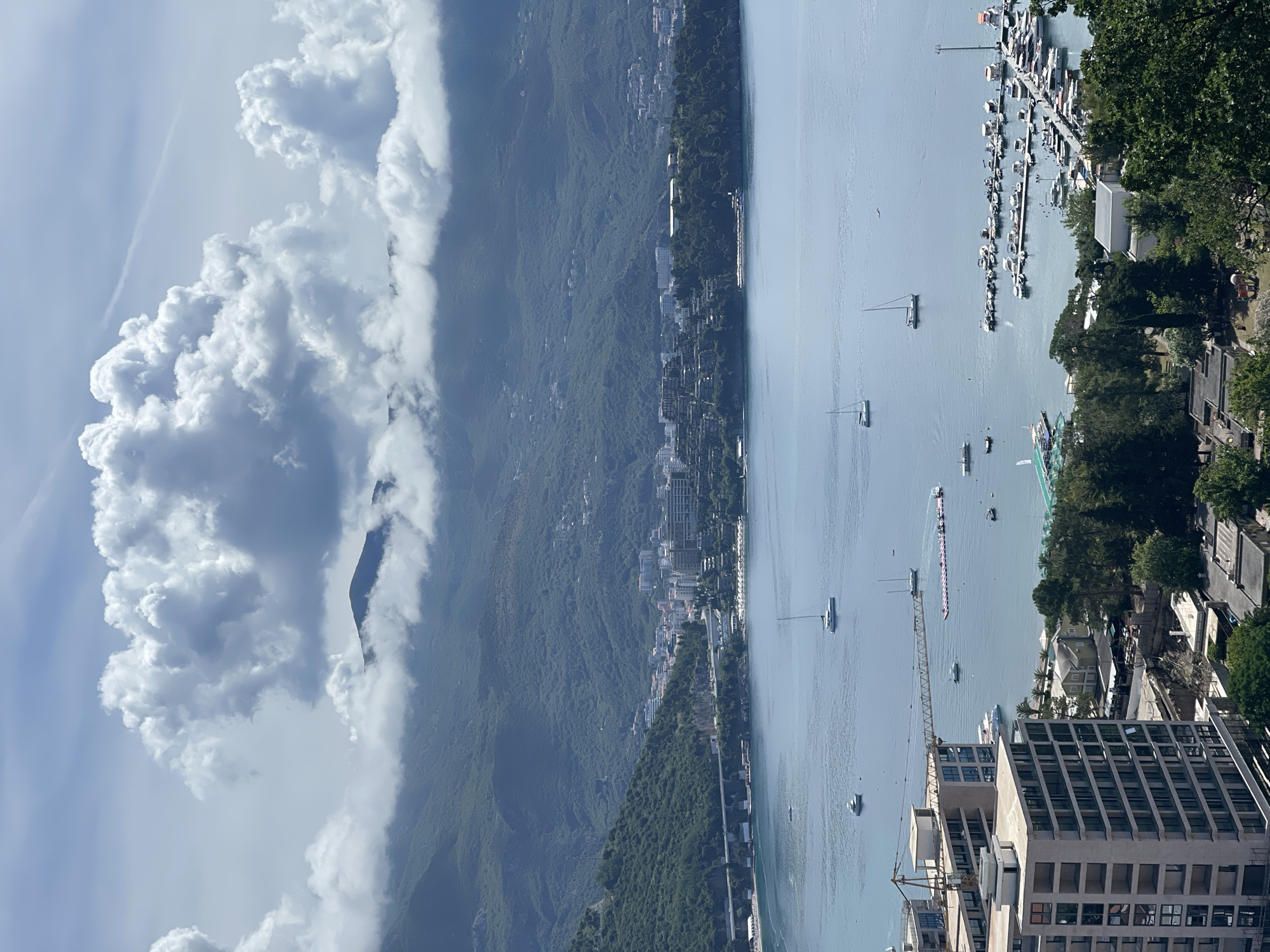 The image displays a wide, panoramic view of the coastline of Budva, Montenegro, featuring a stunning blend of natural landscapes and urban development under a bright, sunny sky.
In the foreground, on the right side of the frame, are several buildings, including a prominent modern structure under construction, indicated by a tall yellow crane. Adjacent to this developed area, a bustling marina stretches into the clear blue water, packed with numerous boats, primarily smaller pleasure craft, yachts, and motorboats, docked in neat rows. Lush green trees are visible along the coastline near the marina.
The expansive body of water, likely the Adriatic Sea or a bay, occupies the midground. It is dotted with several sailboats, some with masts visible against the horizon, and other smaller vessels. A long, narrow, multi-colored floating structure, possibly a buoy line or marker for an event, can also be seen on the water.
Across the bay, on the left side of the image, a dramatic range of verdant, tree-covered mountains rises steeply. Along the base of these mountains, bordering the water, is a continuous stretch of urban landscape, indicating the town of Budva with its buildings and infrastructure nestled into the natural topography.
The sky is a vibrant blue, adorned with large, fluffy white cumulus clouds. These clouds are particularly prominent over and around the mountain peaks, adding depth and shadow to the scene. The bright sunlight suggests the photograph was taken during the day, likely in the morning or early afternoon. No specific individuals are clearly discernible within the frame, nor is any visible text present. The overall impression is one of a picturesque and active coastal environment.
