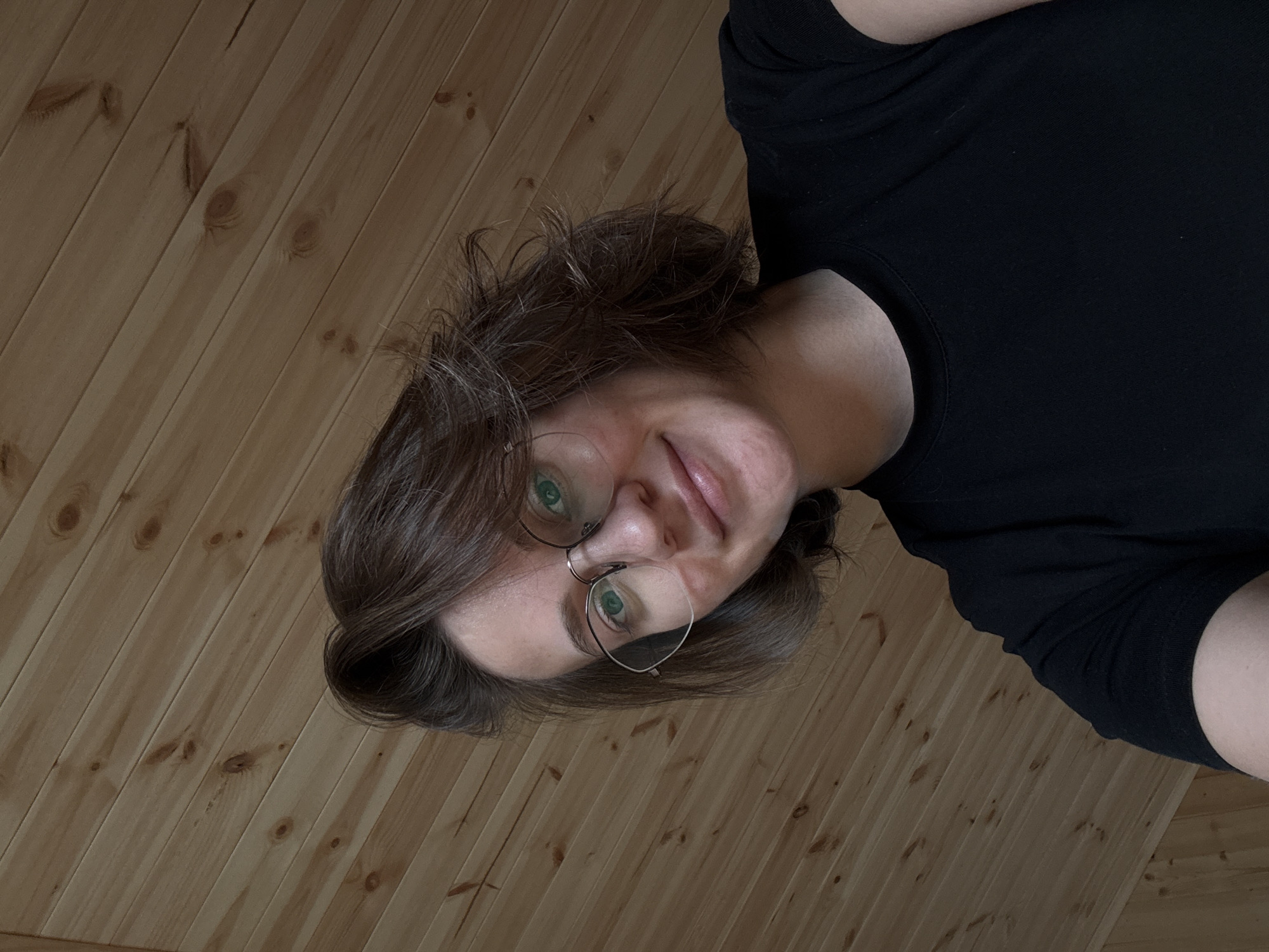 A young adult female is captured in a close-up, low-angle selfie, looking directly at the camera with a faint, closed-mouth smile. She has short, dark brown hair styled with some volume. She wears silver-toned, rounded spectacles, with distinct green reflections visible in the lenses, likely from her eyes. Her skin appears clear, and her lips are naturally colored. She is dressed in a plain black, short-sleeved t-shirt with a rounded neckline. The background consists of light-colored wooden paneling, displaying natural wood grain patterns and knots, oriented diagonally from the upper left to the lower right. The scene is illuminated by a warm, soft light. No specific visual elements within the image provide confirmation of a location in the city of Lenino, Russia.