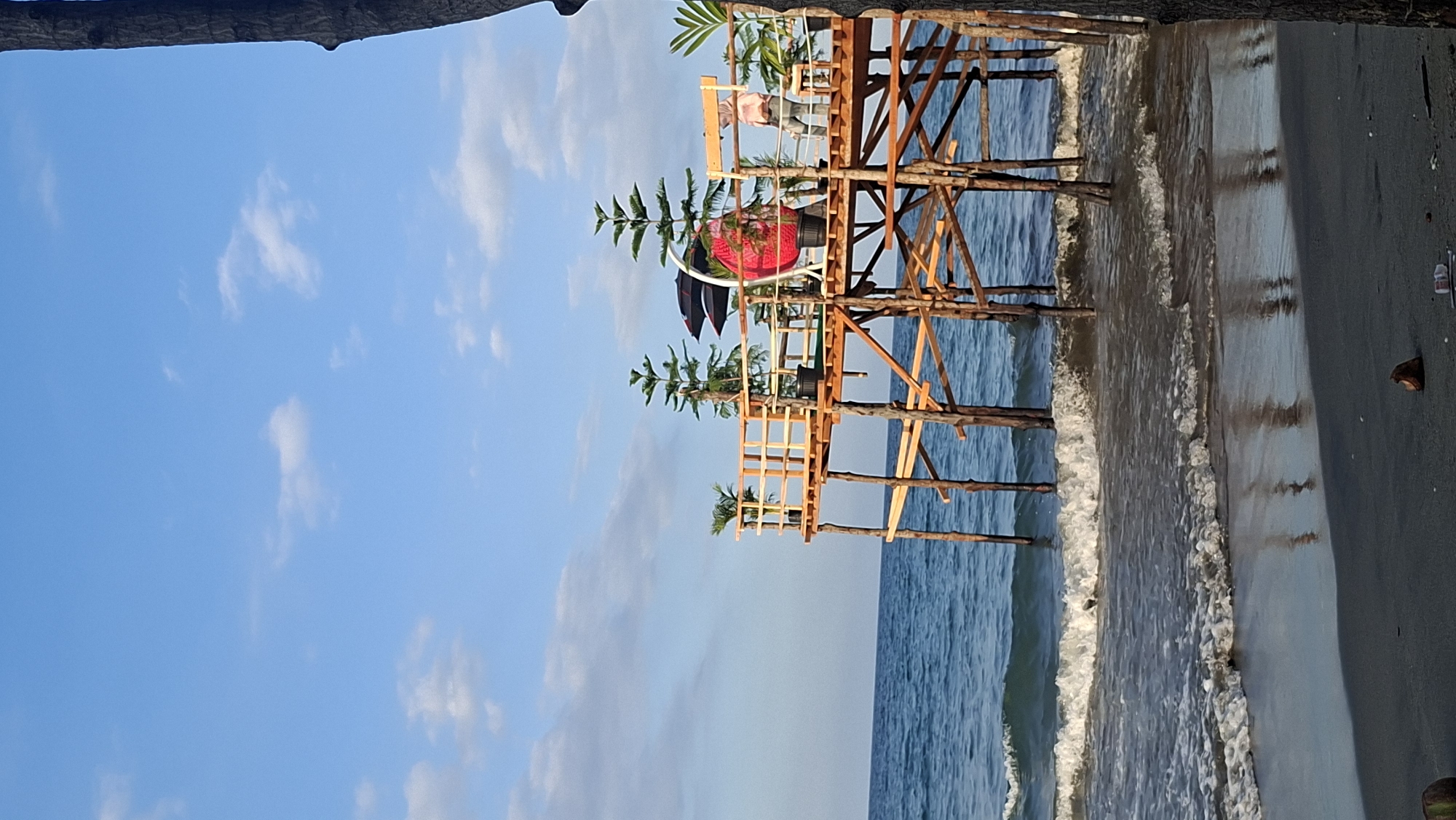 An elevated wooden platform with a railing extends over the ocean at a beach in Lhokseumawe, Indonesia. The platform is supported by numerous wooden posts. Several potted plants, including two tall, narrow pine-like trees and a smaller palm, are placed on the platform. A bright pink hanging chair with a white frame is positioned in the center of the platform. To the right, a person wearing a light-colored headscarf and a pink shirt stands near the railing. The ocean waves are breaking on the sandy shore, with white foam visible. The sky is blue with scattered white clouds. The foreground shows the dark sand beach with a few scattered coconuts and a small white plastic bottle. A large tree trunk is visible on the far right edge of the frame.