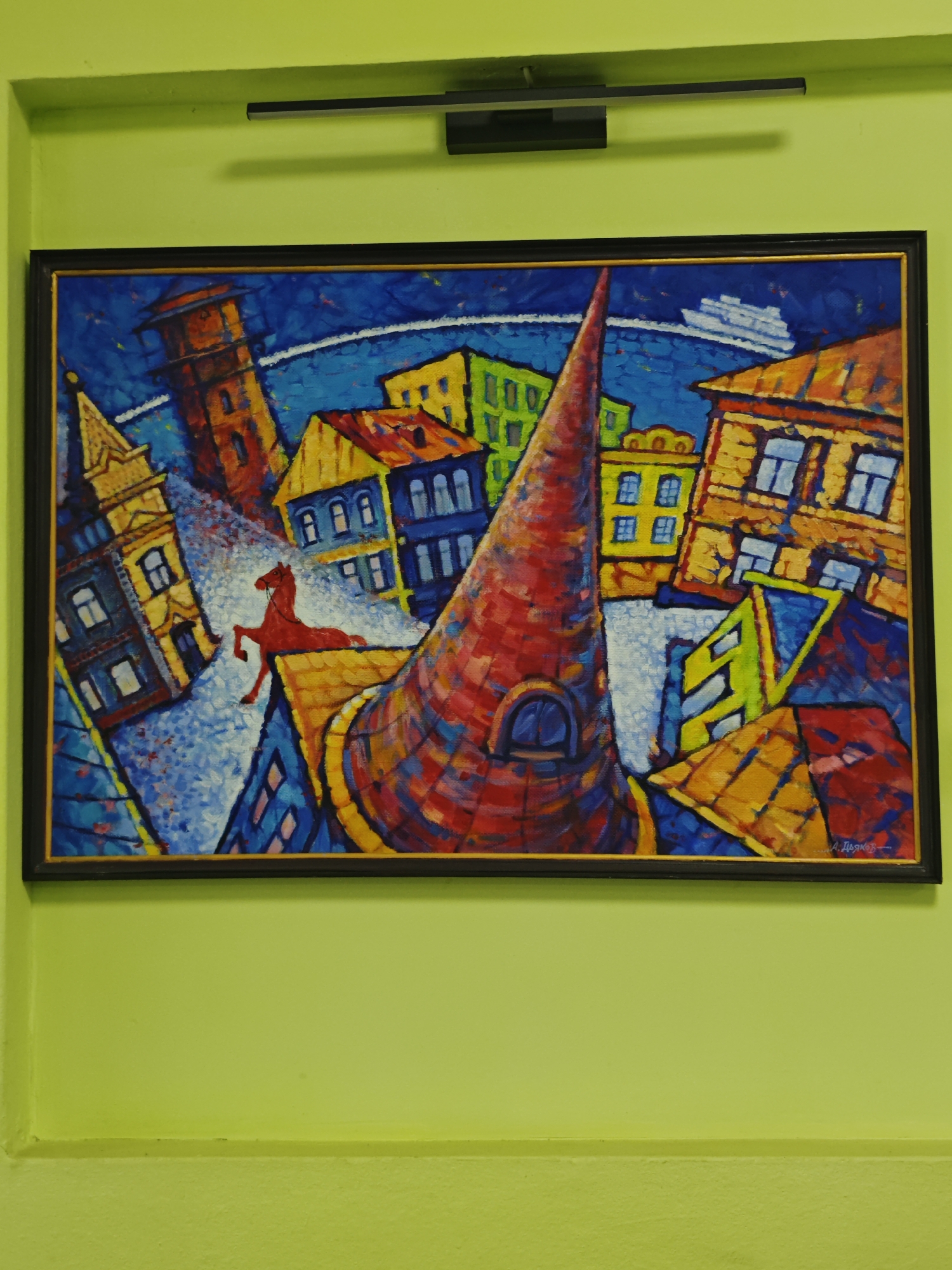 The image shows a framed painting of a city with a red horse in the foreground, set against a bright green wall. The painting depicts a stylized cityscape with colorful buildings in varying shades of yellow, blue, red, and green. A tall, conical red tower dominates the center of the composition, with a red horse positioned on the roof of a yellow building below it. A white ship is visible in the background. The style of the painting is abstract and cubist-inspired. A signature that appears to be "А. Дяков" is present in the bottom right corner of the painting. The painting is lit by a black light fixture mounted above it on the green wall. The wall itself is painted a vibrant lime green and has a subtle textured finish. The painting is oriented horizontally and is the central focus of the image. The city depicted in the painting is not identifiable as Khvalynsk, Russia, based on the visual information provided.