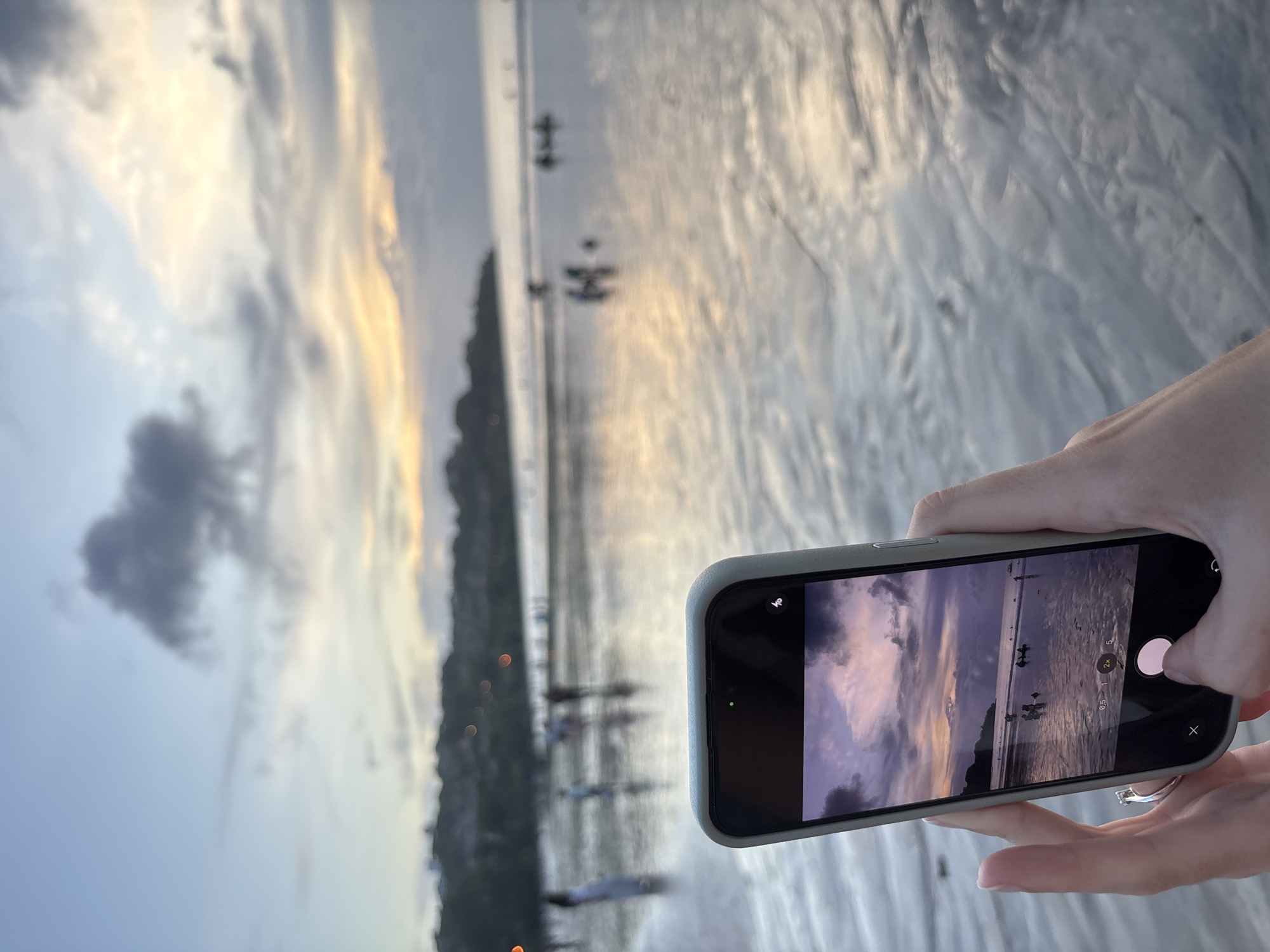 A person's hands hold an iPhone with a light grey case, capturing a serene sunset or sunrise over a coastal landscape. In the foreground, the hands, with a silver ring on the right index finger, grasp the smartphone horizontally. The phone's screen displays a live preview of the scene, confirming the user is taking a photo. Visible camera controls on the screen include zoom options "0.5", "1", and "2x", along with a white circular shutter button. Battery and signal icons are also faintly visible at the top right of the screen.
The smartphone is aimed at a wide, calm body of water, likely a sea or large bay, stretching towards a distant shoreline. The water's surface beautifully reflects the warm, soft light of the setting or rising sun, creating a shimmering effect. Several blurred figures, possibly people wading or small boats, are discernible in the shallow water. The sky above features a blend of light blue and dramatic clouds, illuminated with golden and orange hues from the sun, while a more prominent dark cloud rests on the left. The lighting strongly suggests either dawn or dusk, imbuing the scene with a tranquil ambiance. A blurred landmass with indistinct structures lines the distant horizon. The immediate ground under the hands appears to be wet sand or a smooth, reflective surface. This picturesque moment is likely captured at a beach in Kuta Selatan, Indonesia.