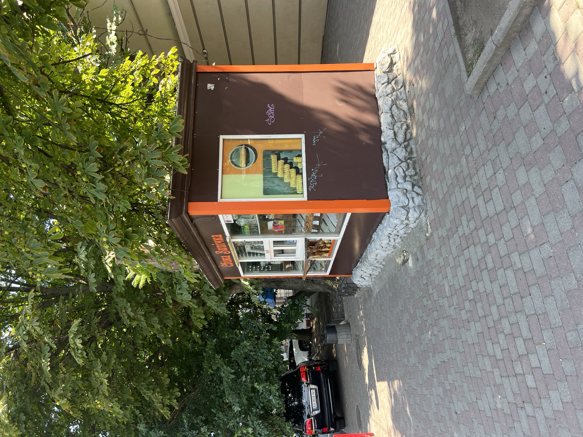 The image shows a small brown kiosk with orange trim, likely a food stand, situated on a brick-paved sidewalk in Odesa, Ukraine. The kiosk features a display window showcasing baked goods, and a smaller window for customer service. Above the service window, text in Cyrillic script, "Свіжа випічка" (Fresh Pastries), is visible. A graphic on the kiosk depicts a spiral of yellow pastries. The kiosk is shaded by a large, leafy tree, and a black Lexus SUV is parked on the street to the left of the kiosk. The background includes a building with beige siding. Graffiti is present on the kiosk's brown side panels. The scene suggests a typical urban setting in Odesa, with a focus on a local business operating outdoors.