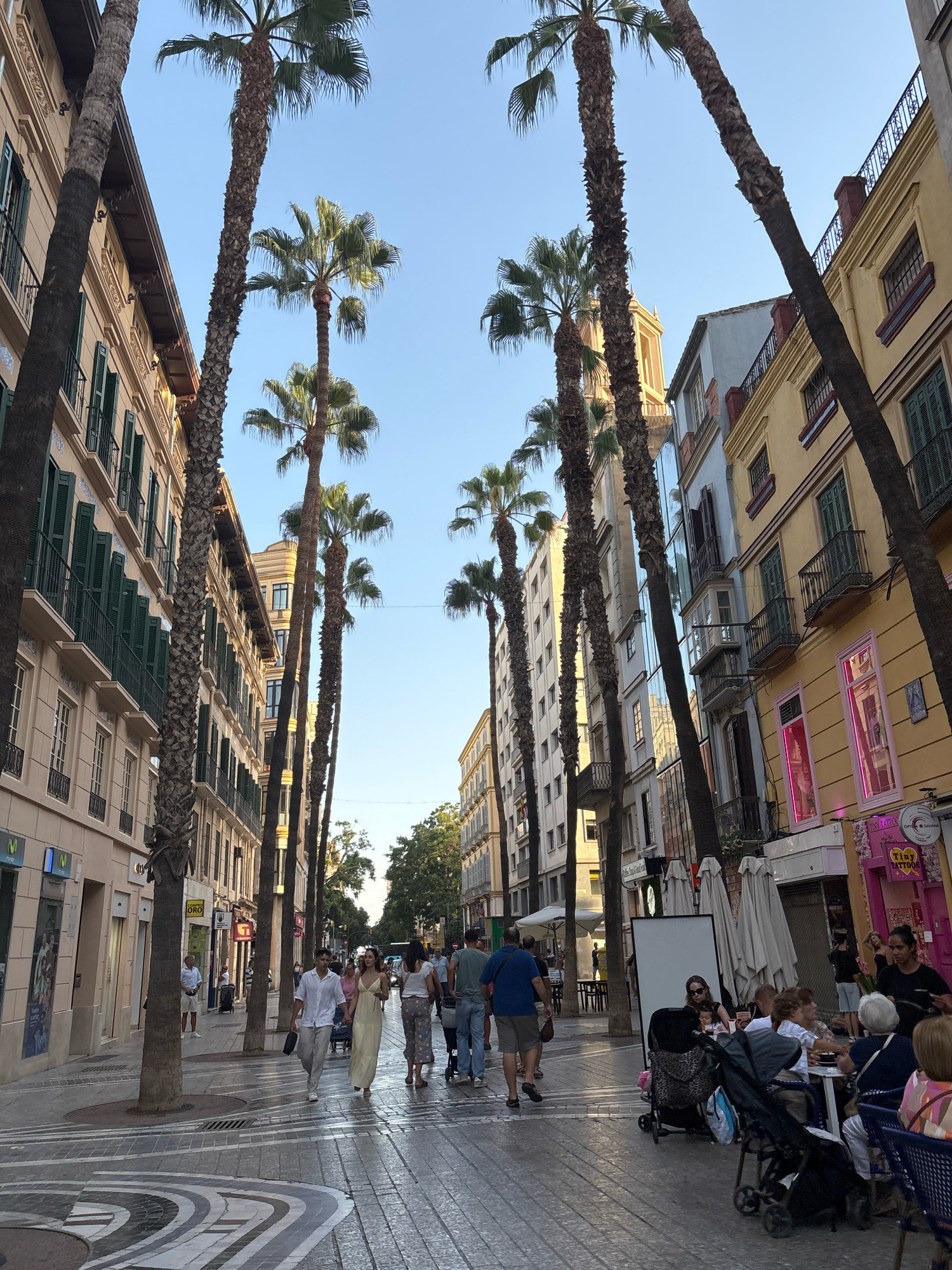 The image depicts a pedestrianized street in Málaga, Spain, likely in the historic city center given the architecture and palm trees. The street is lined with multi-story buildings, featuring balconies and shuttered windows, characteristic of Spanish colonial or Mediterranean design. The buildings display various pastel colors, predominantly yellow and beige, with some showing signs of age. Tall, slender palm trees are planted along the street, their fronds reaching towards a clear blue sky.
Several people are visible walking and seated at outdoor cafes. A couple, the man in a white shirt and light trousers, and the woman in a flowing yellow dress, are walking hand-in-hand towards the camera. Other pedestrians are scattered along the street, some with shopping bags or pushing strollers. A group of people are seated at tables outside a cafe on the right side of the image, engaged in conversation. One individual, a woman with dark hair and sunglasses, is seated next to a stroller.
The paving stones of the street are laid in a patterned design, with a mosaic-like effect. Shops and businesses are integrated into the ground floors of the buildings, with some signage visible, including a pink storefront with neon lighting. The overall atmosphere suggests a lively and pleasant urban environment during daylight hours.