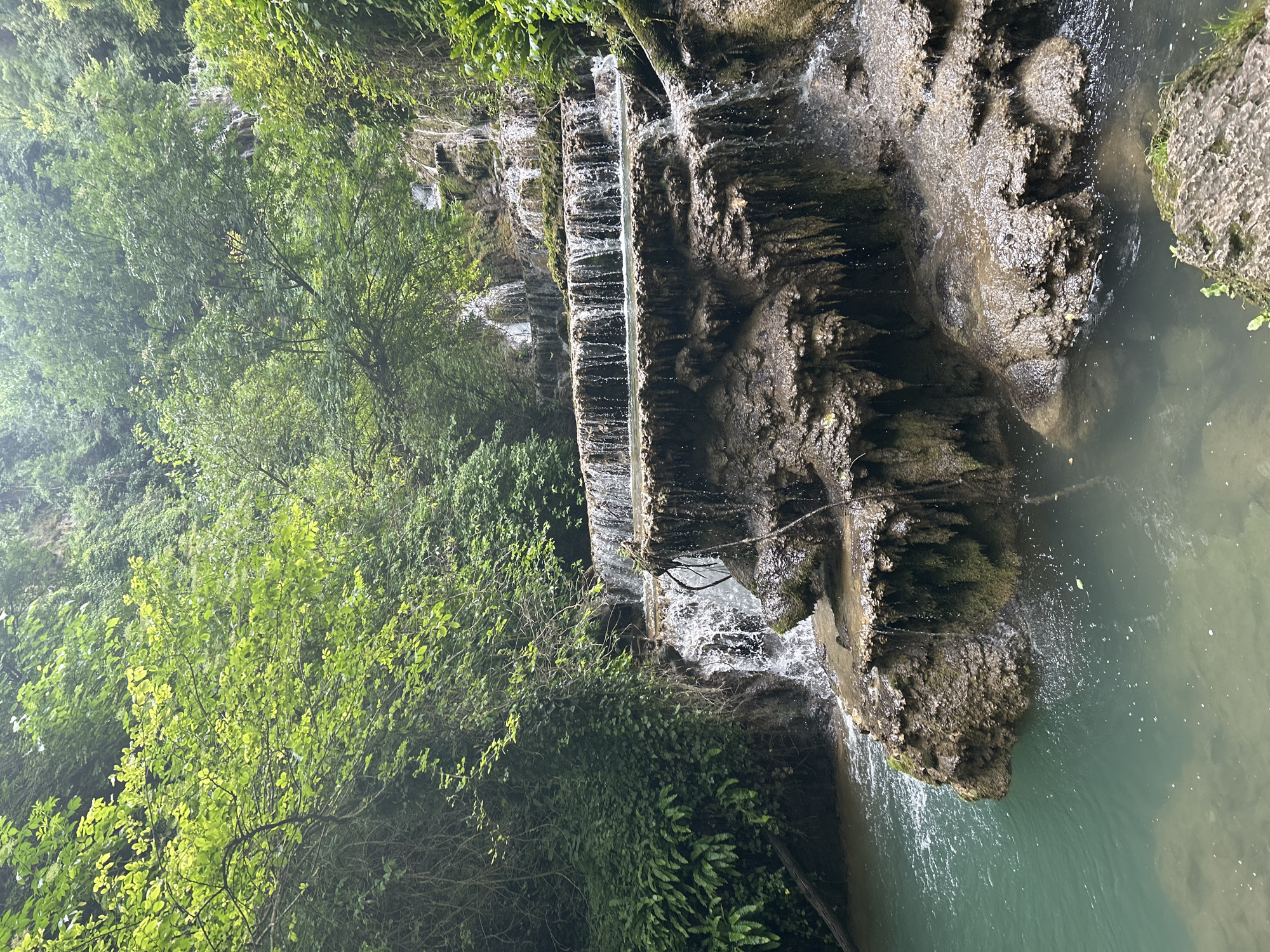 This image depicts the Krushuna Waterfalls in Krushuna, Bulgaria. A multi-tiered waterfall cascades over travertine formations, surrounded by lush green vegetation. The water is a vibrant turquoise and flows into a pool at the bottom. The rocks are wet and mossy, with stalactite-like formations hanging from the ledges. The overall scene is one of natural beauty and tranquility.