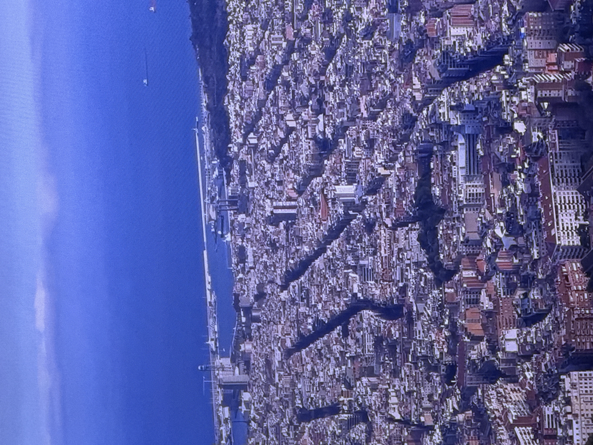 This is an aerial, high-angle, slightly blurry photograph showcasing a dense urban cityscape with a prominent harbor and the ocean in the background. The city is characterized by a grid-like street layout with numerous multi-story buildings clustered tightly together. The architecture appears to be a mix of styles, with many buildings having pitched red-tiled roofs. The streets themselves are narrow and appear to be dark, possibly due to shadows cast by the buildings or the angle of the sun. In the distance, a large harbor area is visible, with docks, cranes, and several vessels. Beyond the harbor lies a vast expanse of deep blue ocean, with a few distant ships and a faint line of clouds on the horizon. The overall impression is one of a bustling coastal city. The image does not depict Dubai, United Arab Emirates.