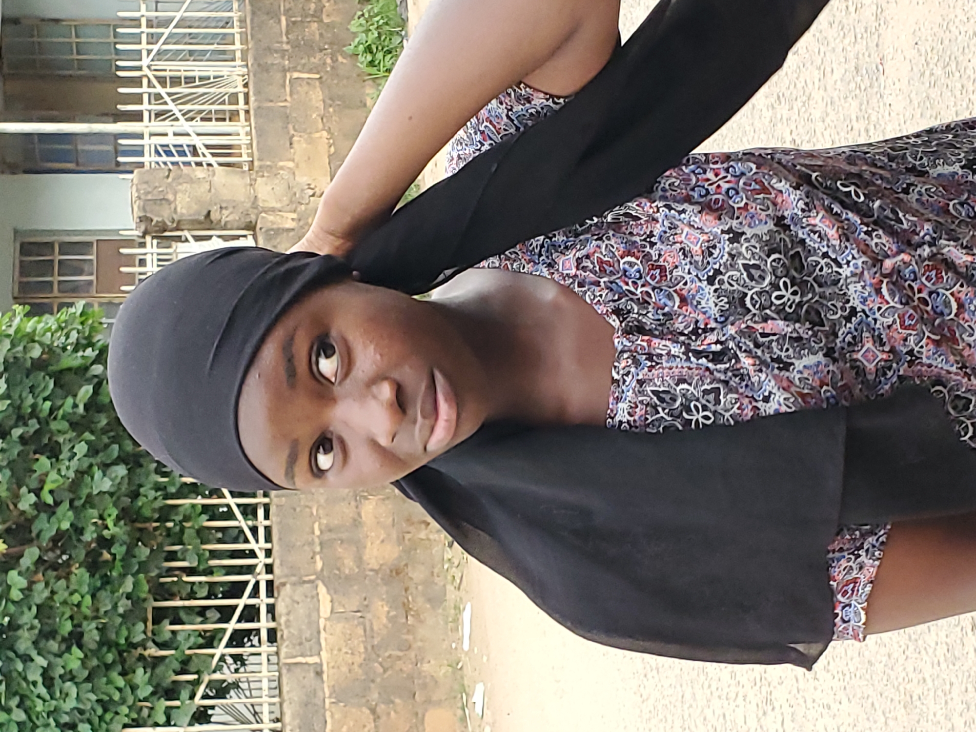 A medium shot captures a woman in Jos, Nigeria. She is wearing a black headscarf and a colorful patterned dress. She looks directly at the camera. A building with white metal bars and green foliage appear in the background. The ground appears to be concrete.