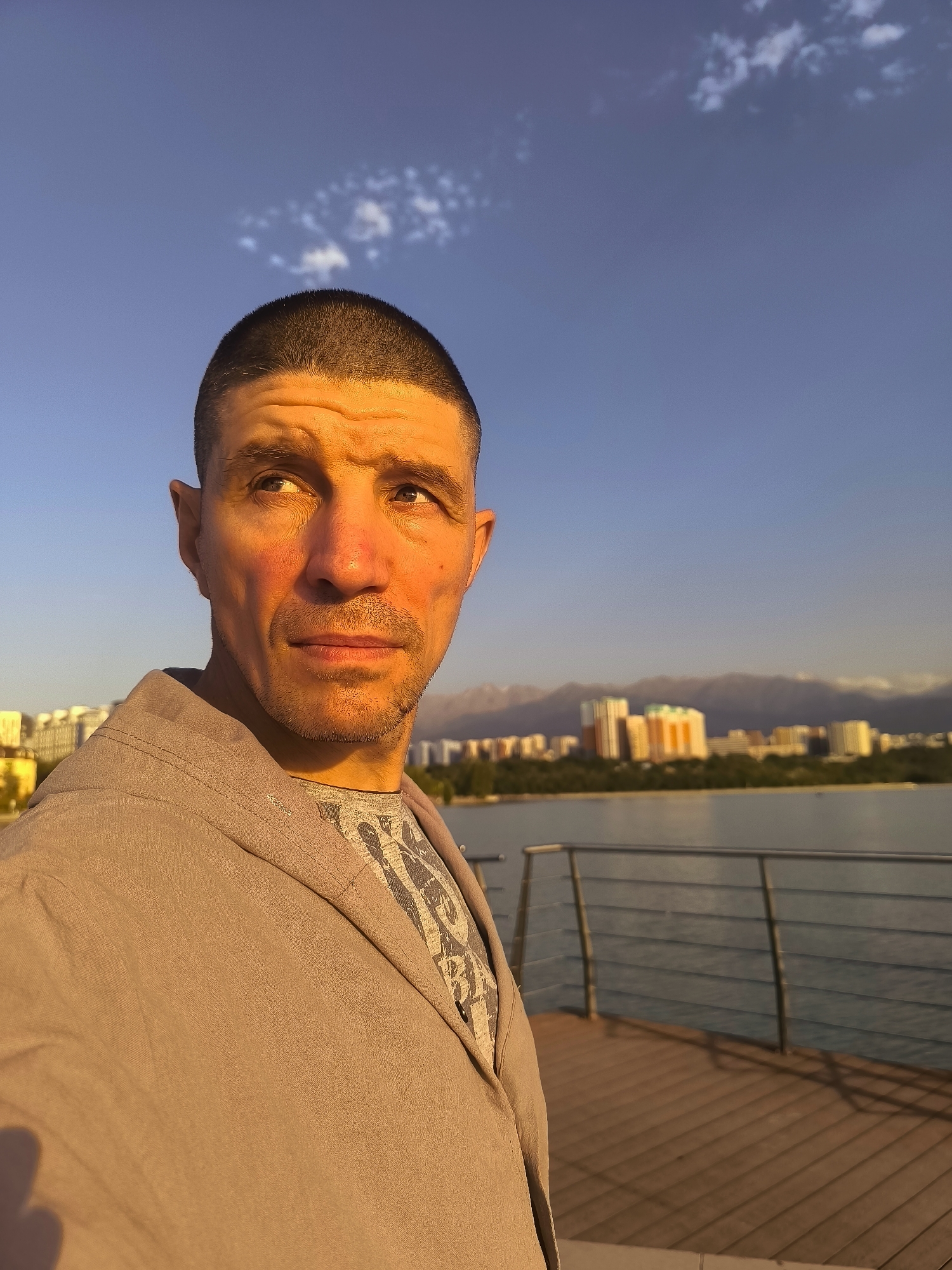 A medium shot shows a man with a buzz cut, wearing a grey jacket over a grey t-shirt, standing on a wooden deck. He is looking to his right with a neutral expression. In the background, a large body of water is visible, with a city skyline and mountains in the distance. The lighting suggests it is late afternoon or early evening, with warm sunlight illuminating the man's face. The scene is set at a waterfront promenade, likely in Almaty, Kazakhstan, possibly near the Big Almaty Lake or another reservoir within or near the city, given the presence of the mountains and urban development.