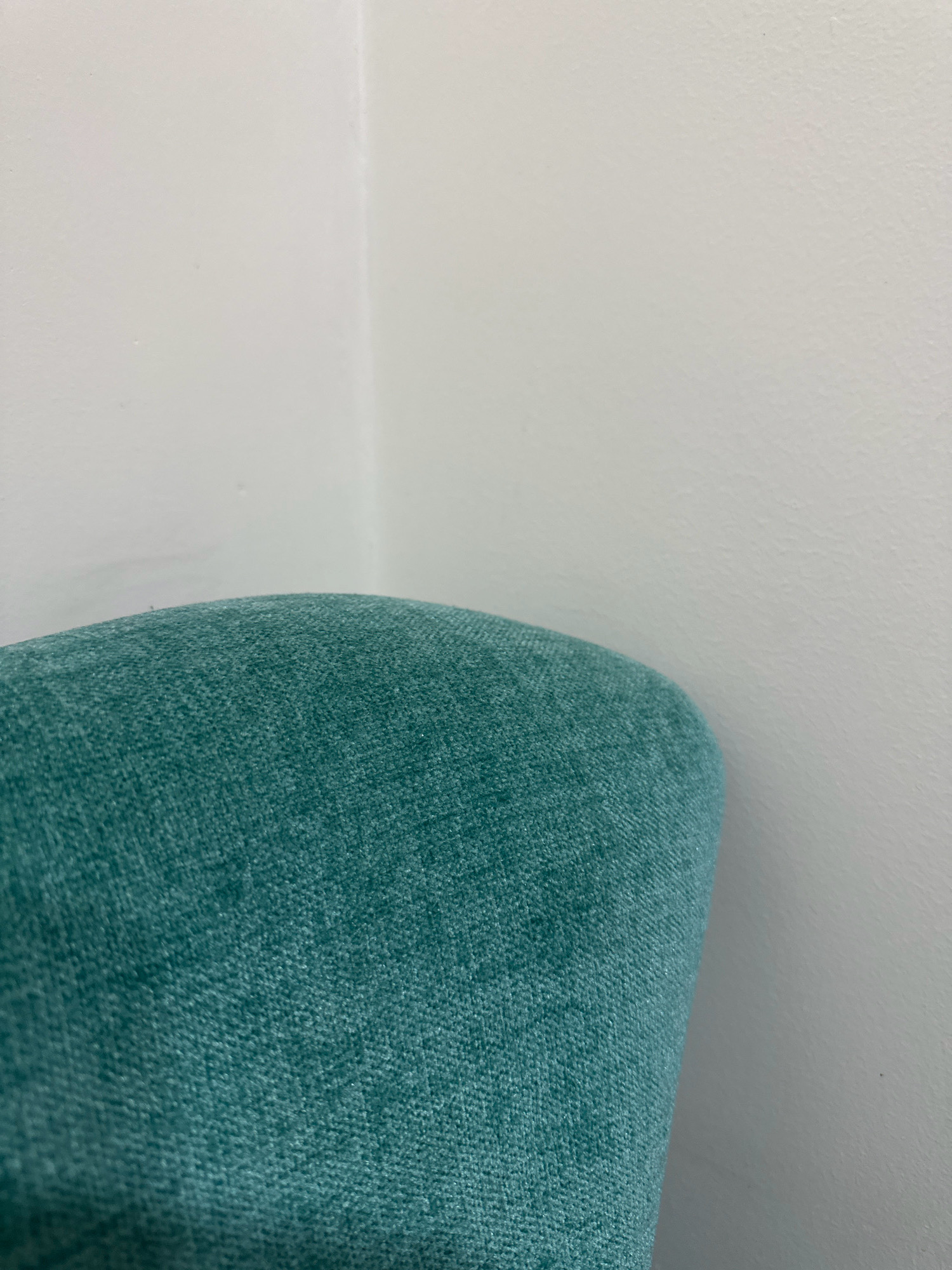 The image shows a close-up, low-angle shot of a teal-colored, textured fabric, likely upholstery, curving upwards and to the right. The fabric fills the bottom half of the frame and appears to be a cushion or part of a piece of furniture. The upper half of the image is dominated by a plain, white wall that meets another white wall at a corner. There are no people, animals, or other objects visible in the scene. The lighting is soft and diffused, creating subtle shadows and highlighting the texture of the fabric. The environment appears to be indoors, in a clean and minimalist setting. This image does not provide any information about the city of Lalitpur, Nepal, or any specific location.