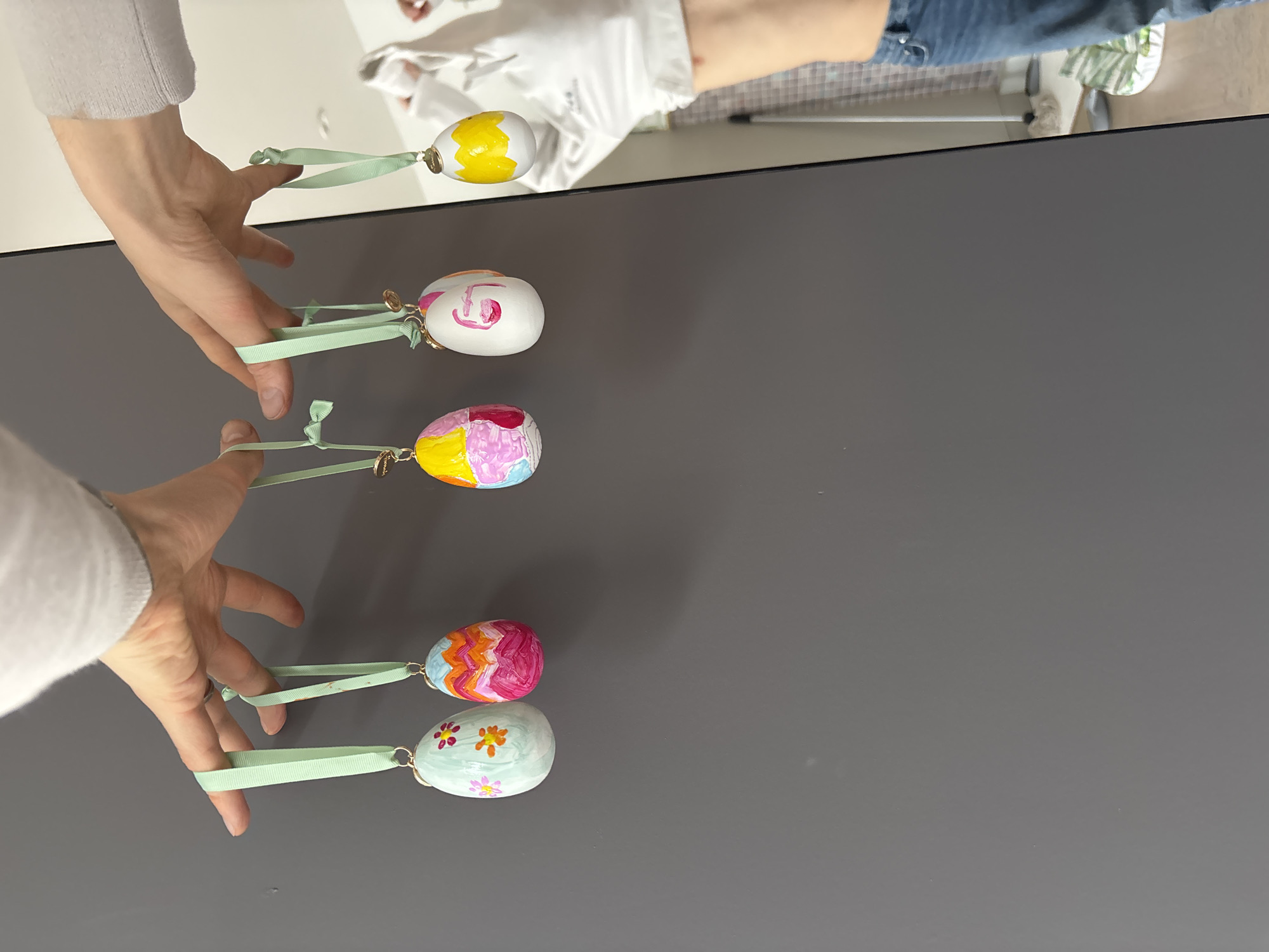 A top-down shot depicts a person holding five painted Easter eggs by their green ribbon loops. The eggs are arranged in a vertical row on a grey surface. Each egg has a unique painted design. The upper-left corner of the image shows parts of the person's arm and hand, clad in a light-colored sweater or sleeve. The eggs are suspended, and the focus is on the eggs and hands against the background surface. The scene appears to be indoors. There is no visible indication that the location is in London, United Kingdom.