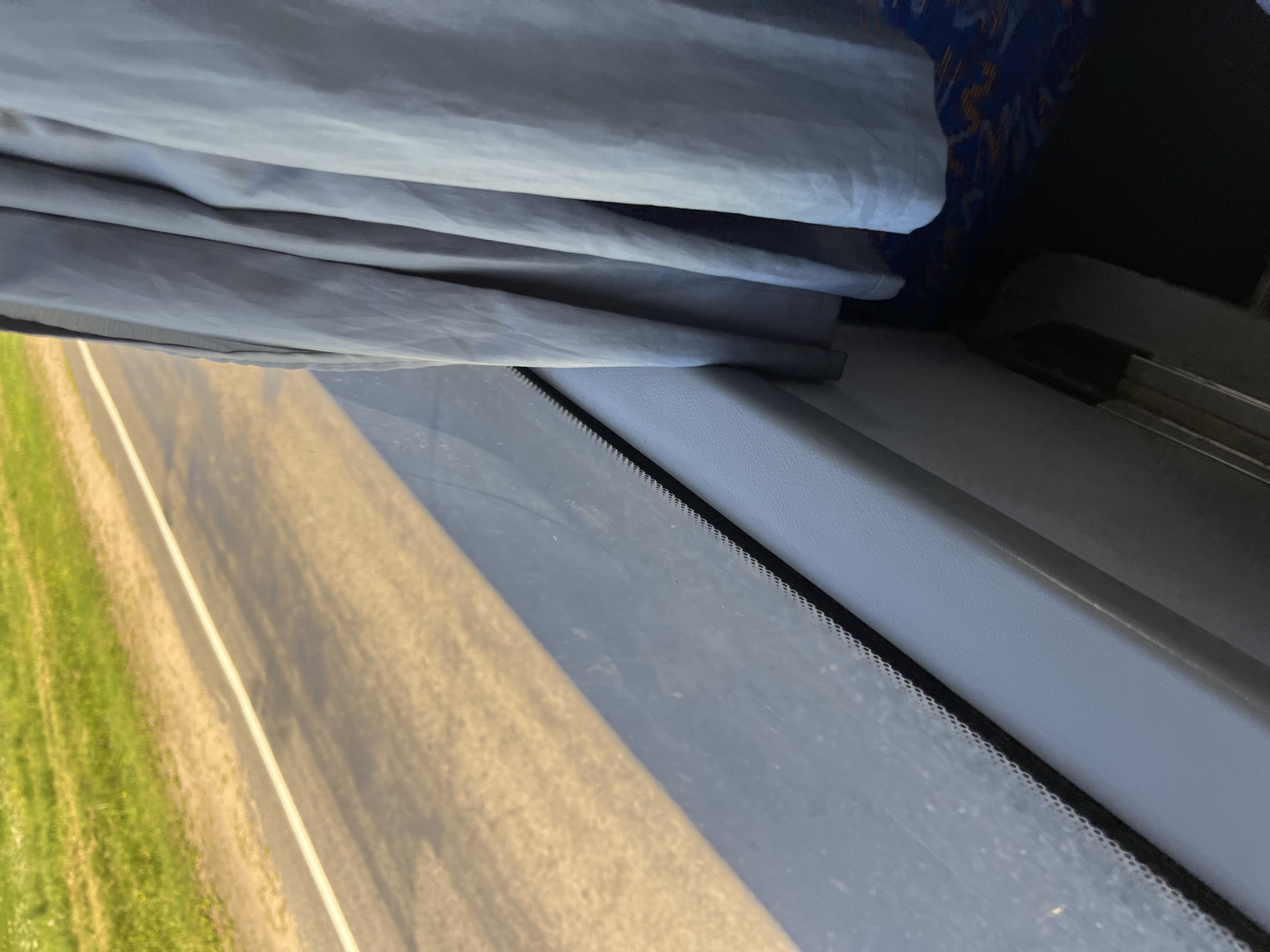 The image shows a view from inside a vehicle, likely a bus, looking out a window. A light blue curtain is pulled partially closed on the right side of the frame. Outside the window, a paved road with a white line is visible, bordered by a grassy area. The sun is casting shadows across the road, indicating it is daytime. The interior of the vehicle shows a gray surface beneath the window, with a patterned seat visible in the lower right corner. There are no people visible in the image. The scene suggests travel through a rural or suburban area. The location is identified as the city of Myadzyel, Belarus.