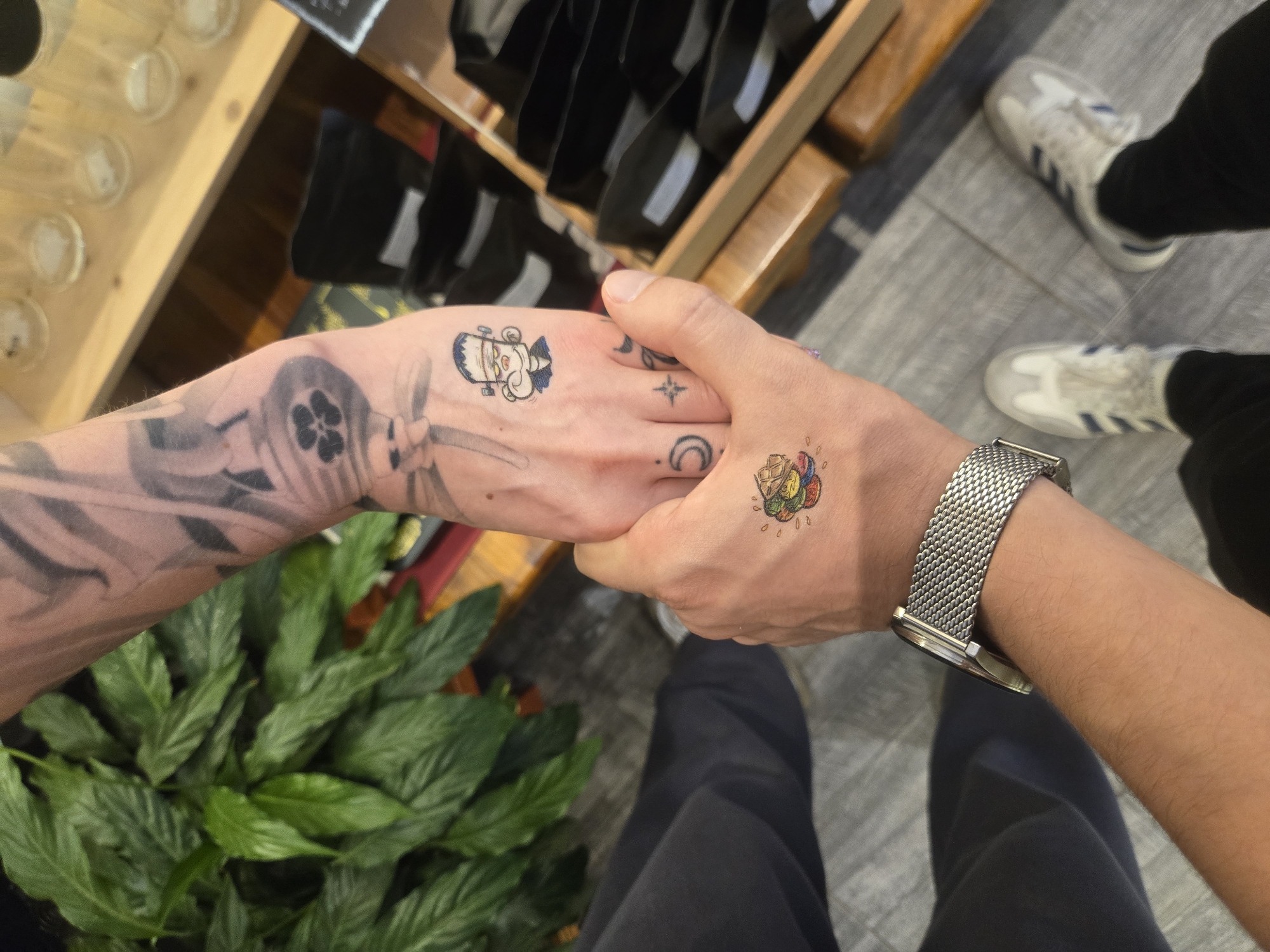 Two people hold hands. The person on the left has a tattoo sleeve. The person on the right has a watch. Both people have cartoon tattoos on their hands. The location appears to be indoors, with a plant and shelves visible. There is no indication in the image that this photo was taken in the city of Budva, Montenegro.